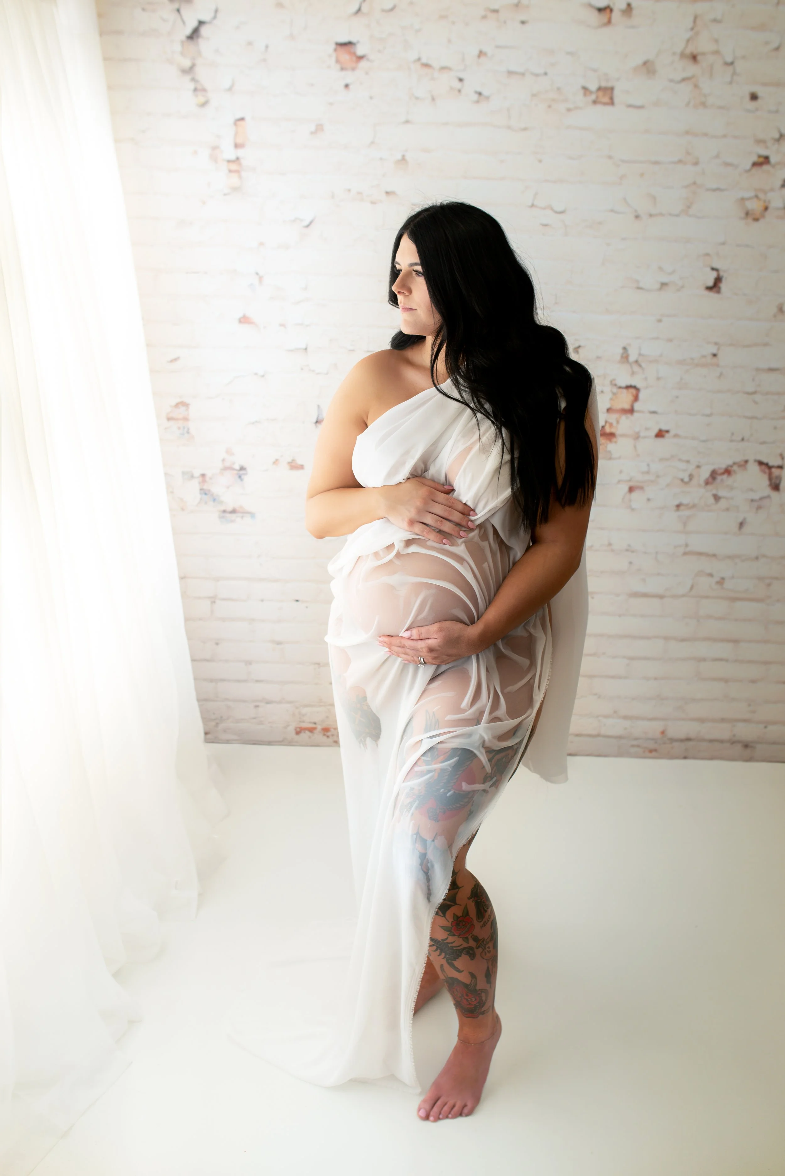 A pregnant woman with long black hair and tattoos on her leg and arm, standing indoors near a white brick wall and sheer curtains, wearing a semi-transparent white dress.