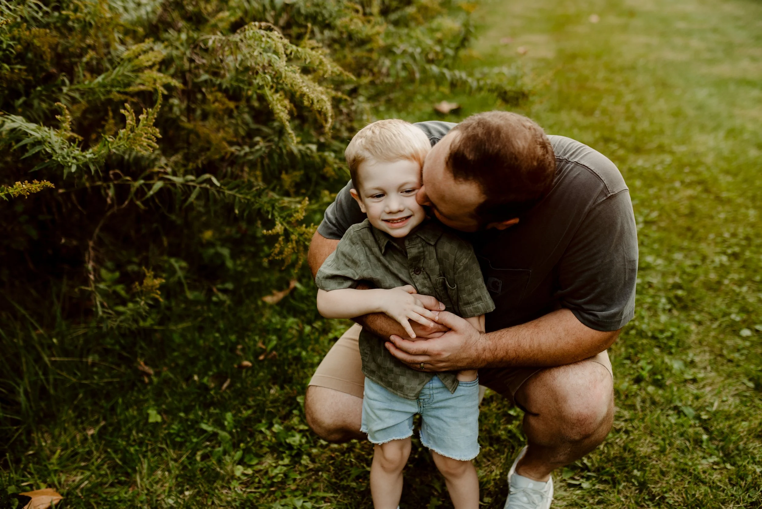 A man hugging and kissing a young boy outdoors on a grassy area, with greenery in the background.