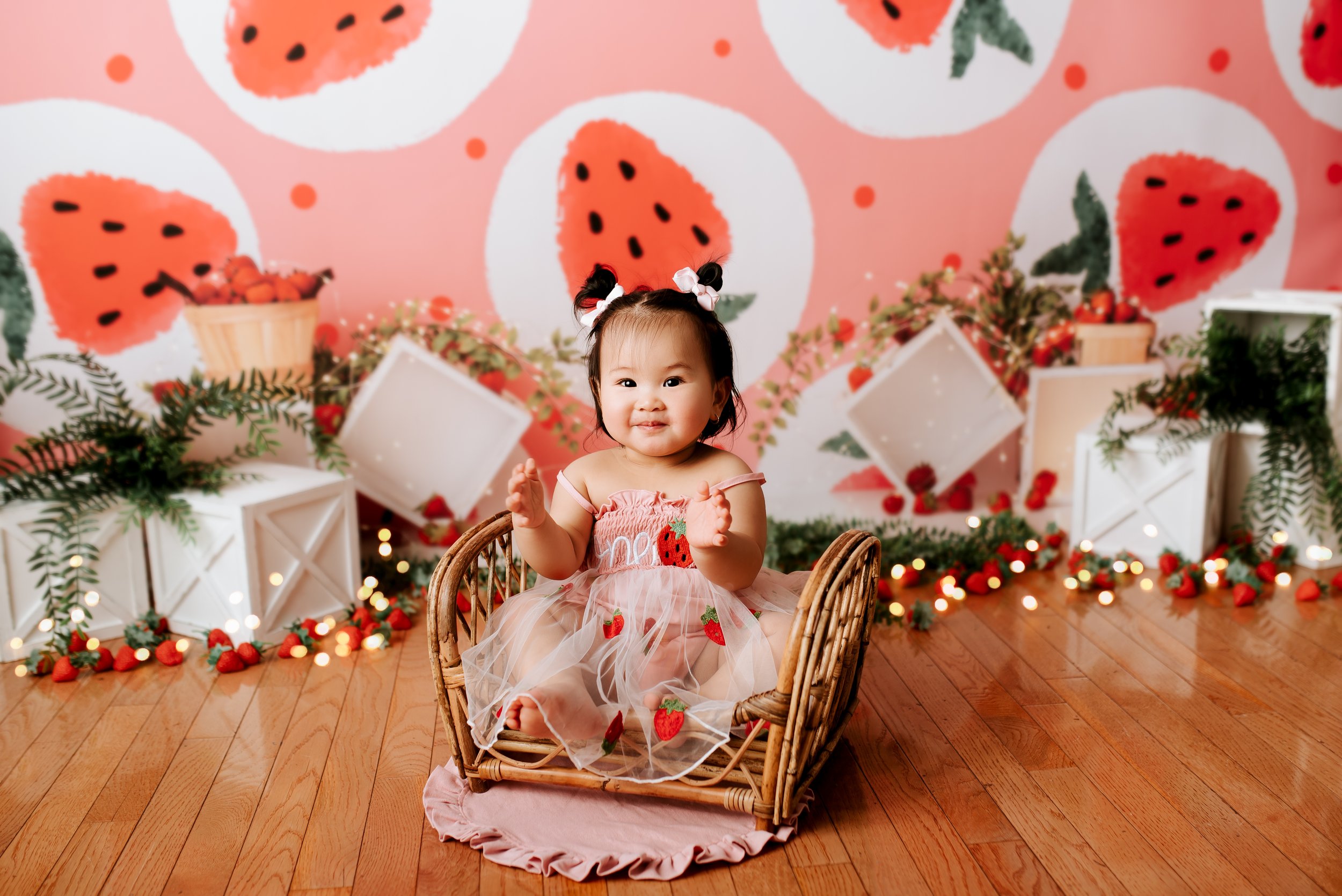 A smiling baby girl in a pink strawberry dress sitting in a wicker basket in front of a pink watermelon-themed backdrop with strawberries, green foliage, and fairy lights.