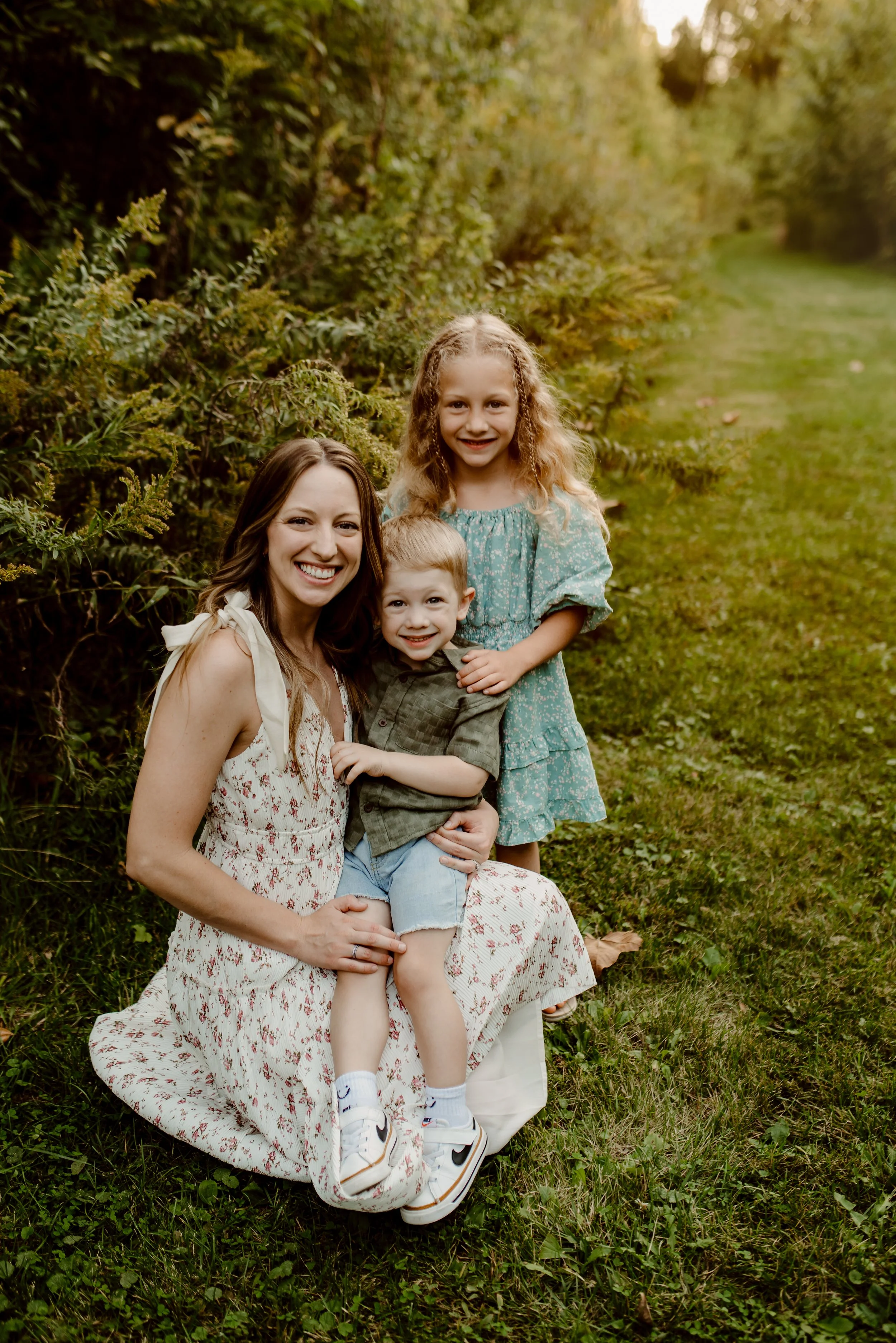 A woman with two children on a grassy trail surrounded by trees, smiling at the camera.