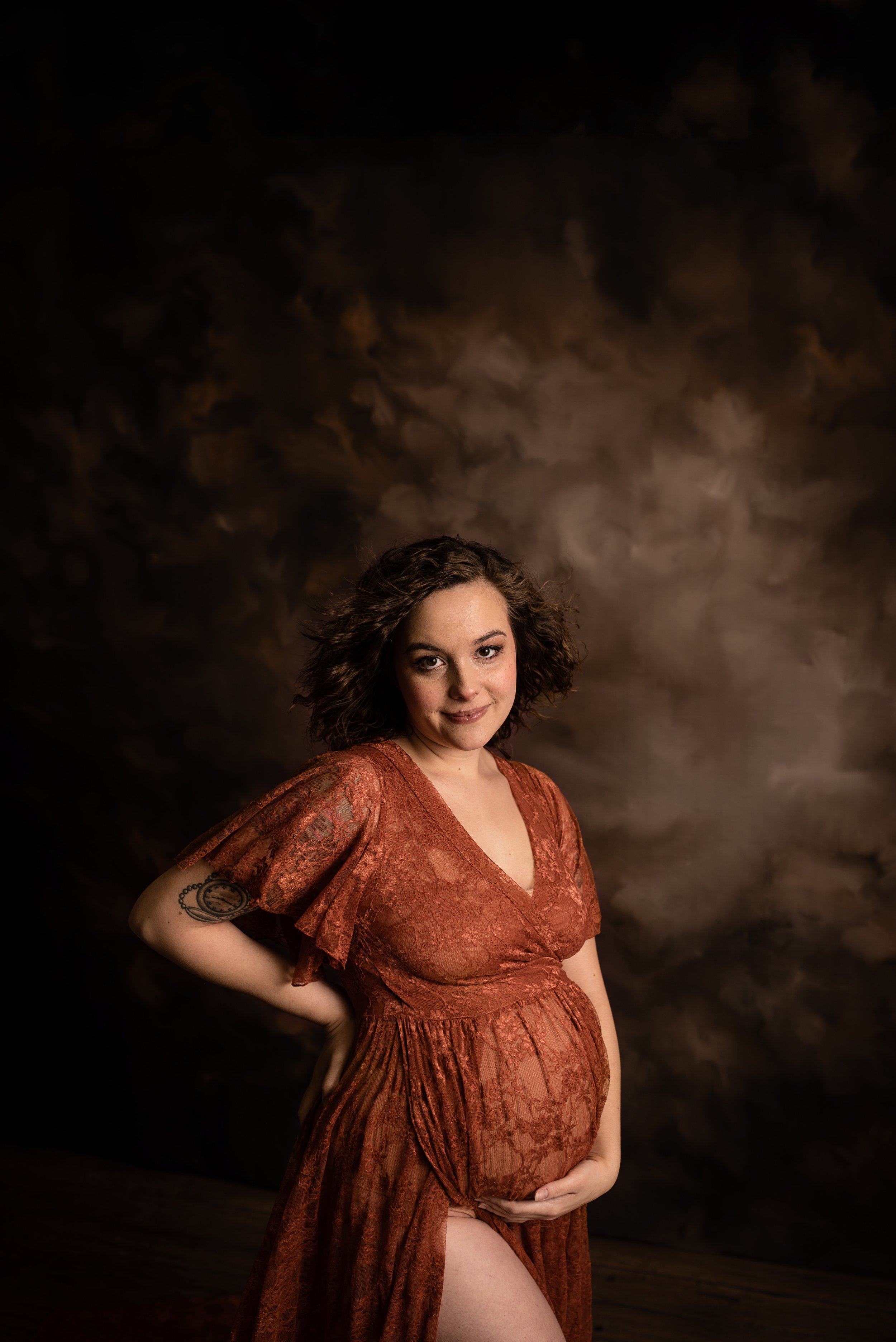 A pregnant woman with curly brown hair wearing a rust-colored lace dress poses with one hand on her hip and the other cradling her belly, standing against a dark, moody cloudy backdrop.