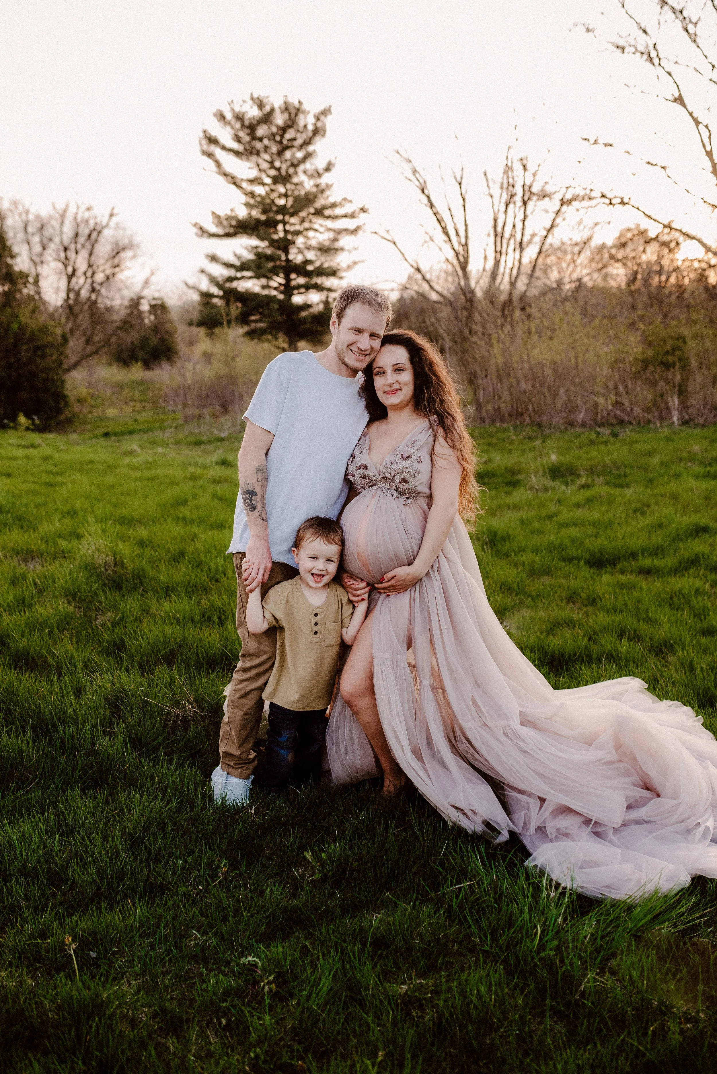 A family of three standing outdoors on green grass during sunset, with trees in the background. The pregnant woman in a flowing pink dress is holding her belly, smiling. The man and young boy are standing beside her, also smiling and holding hands.