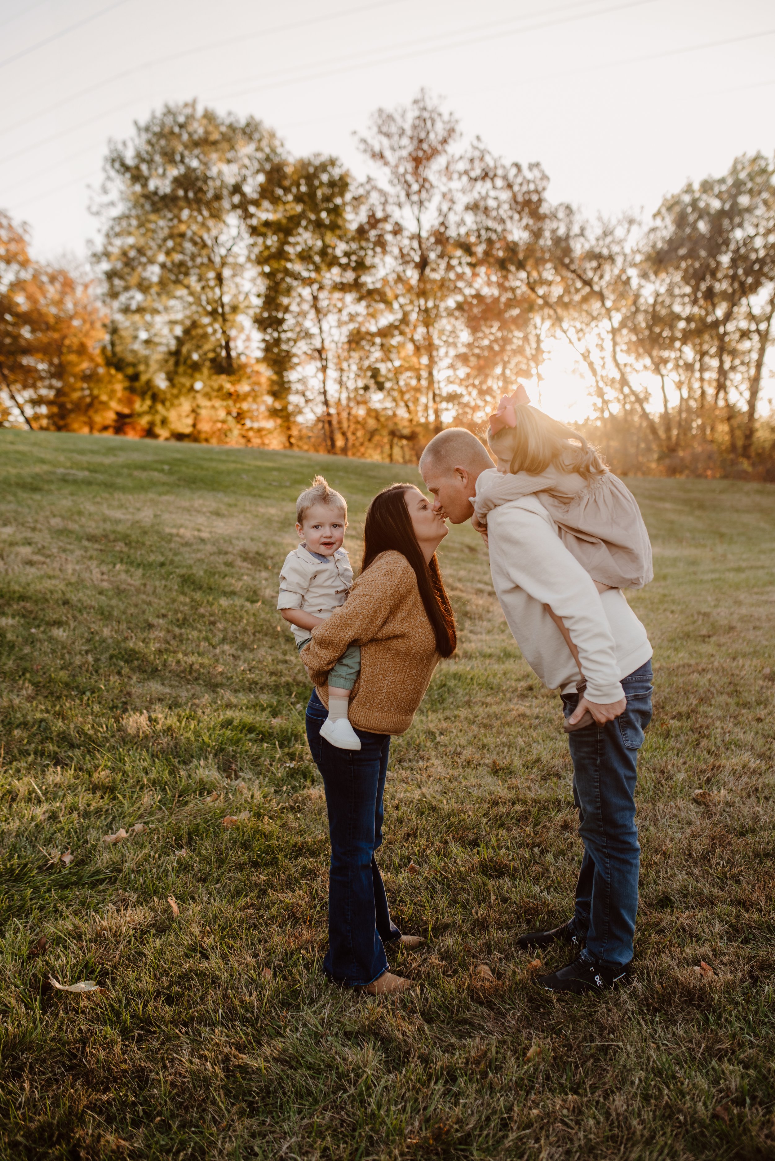 A family of four on a grassy hill during sunset, with two parents kissing and their two young children, one on each parent's back.
