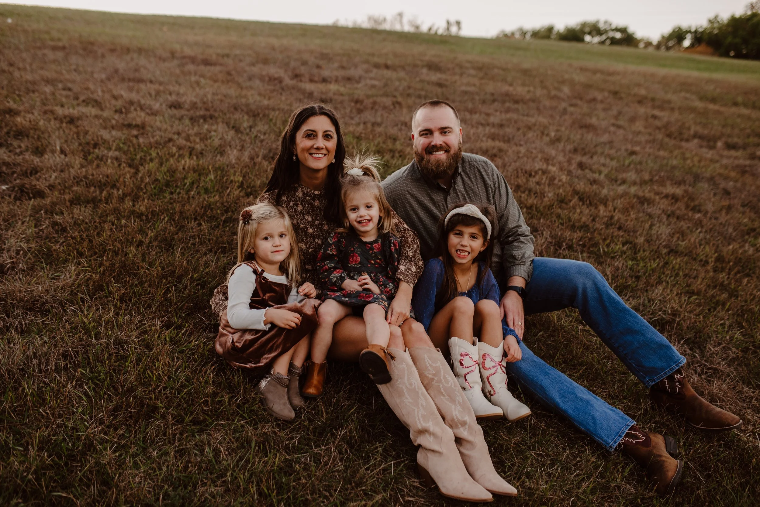 Family of five sitting on a grassy field, smiling at the camera.