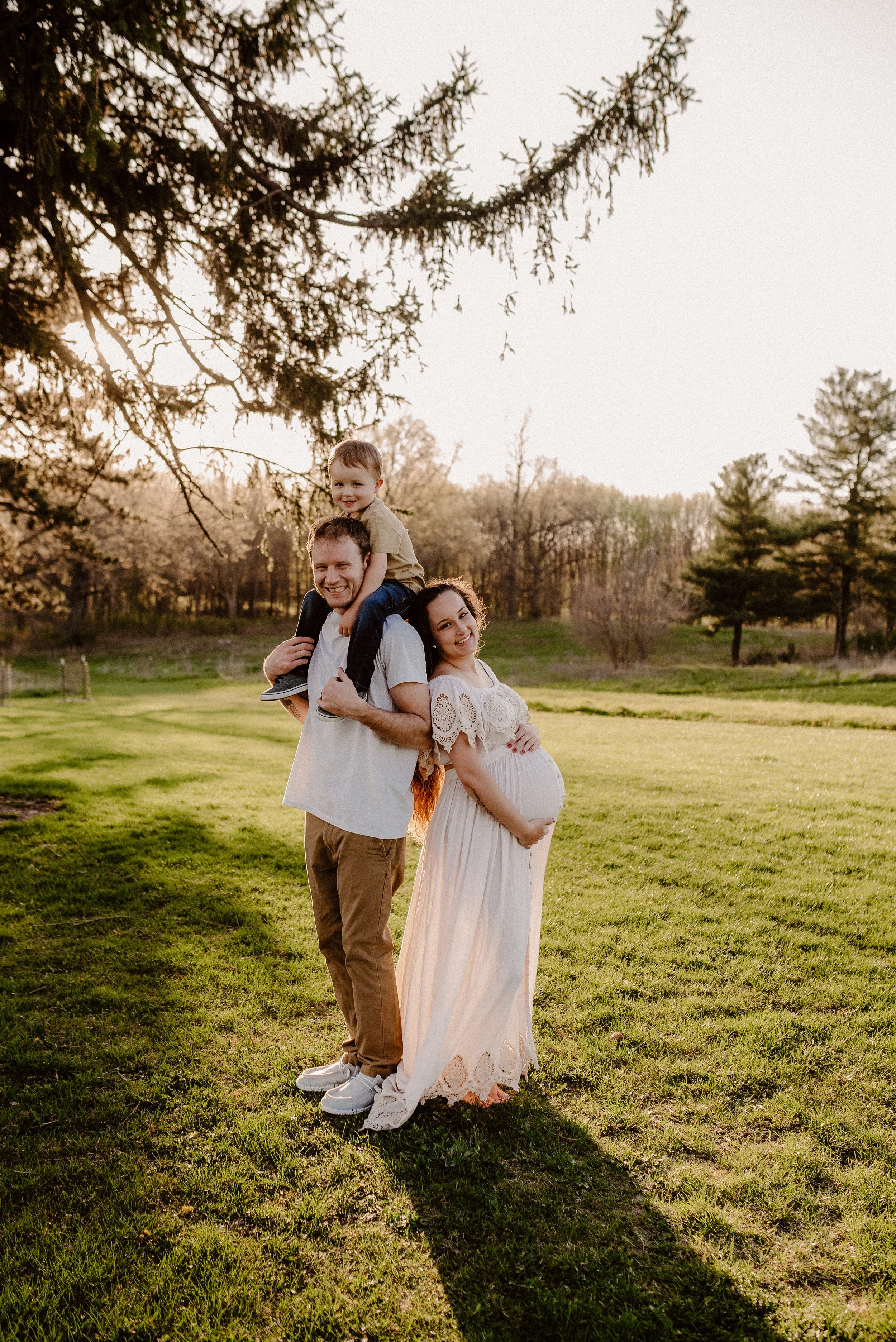 A happy family of three, including a pregnant woman, a man, and a young boy, outdoors on a sunny day with green grass and trees in the background.
