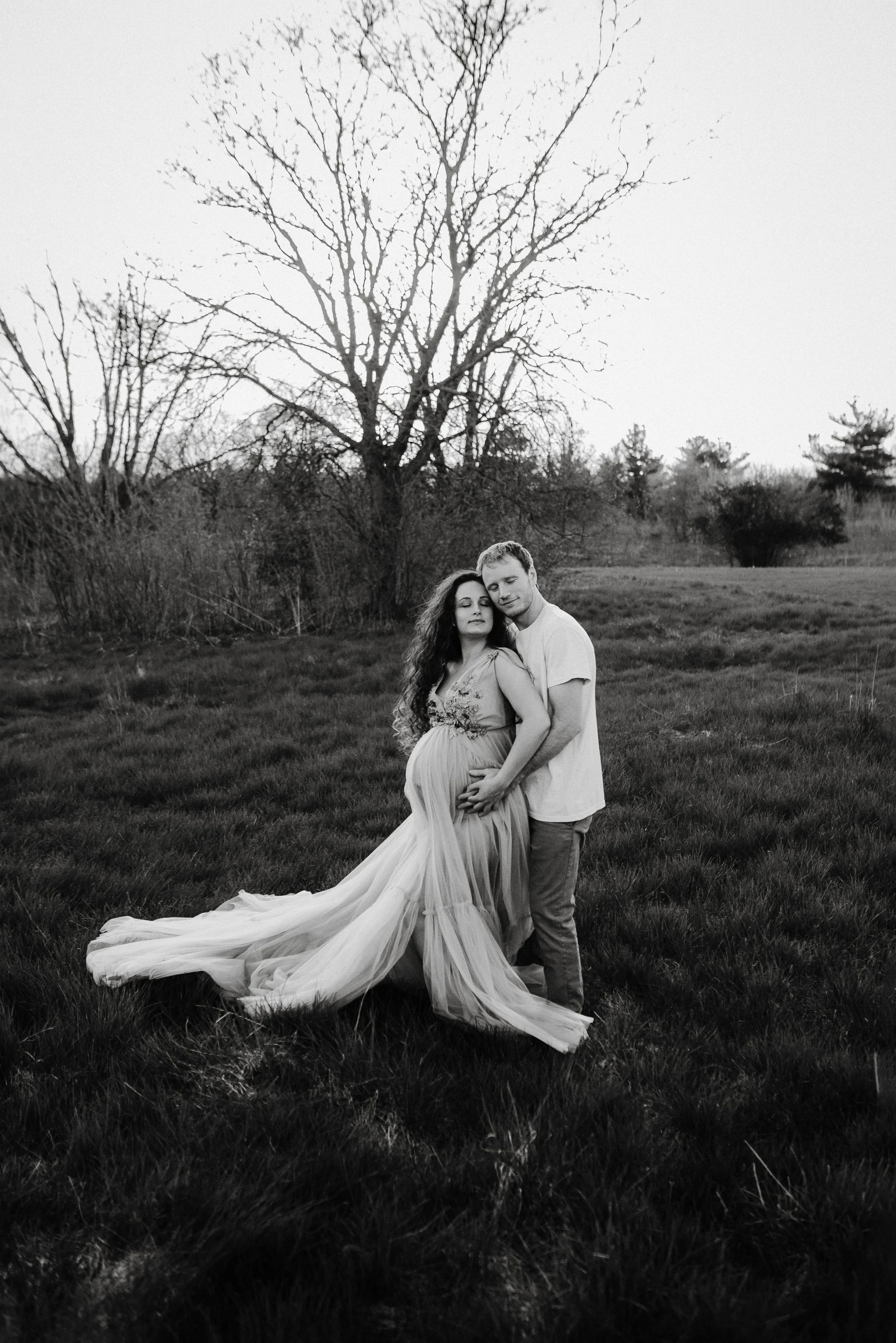 A black and white photo of a pregnant woman in a flowing dress and a man standing behind her with his arms around her in a field with trees and grass.