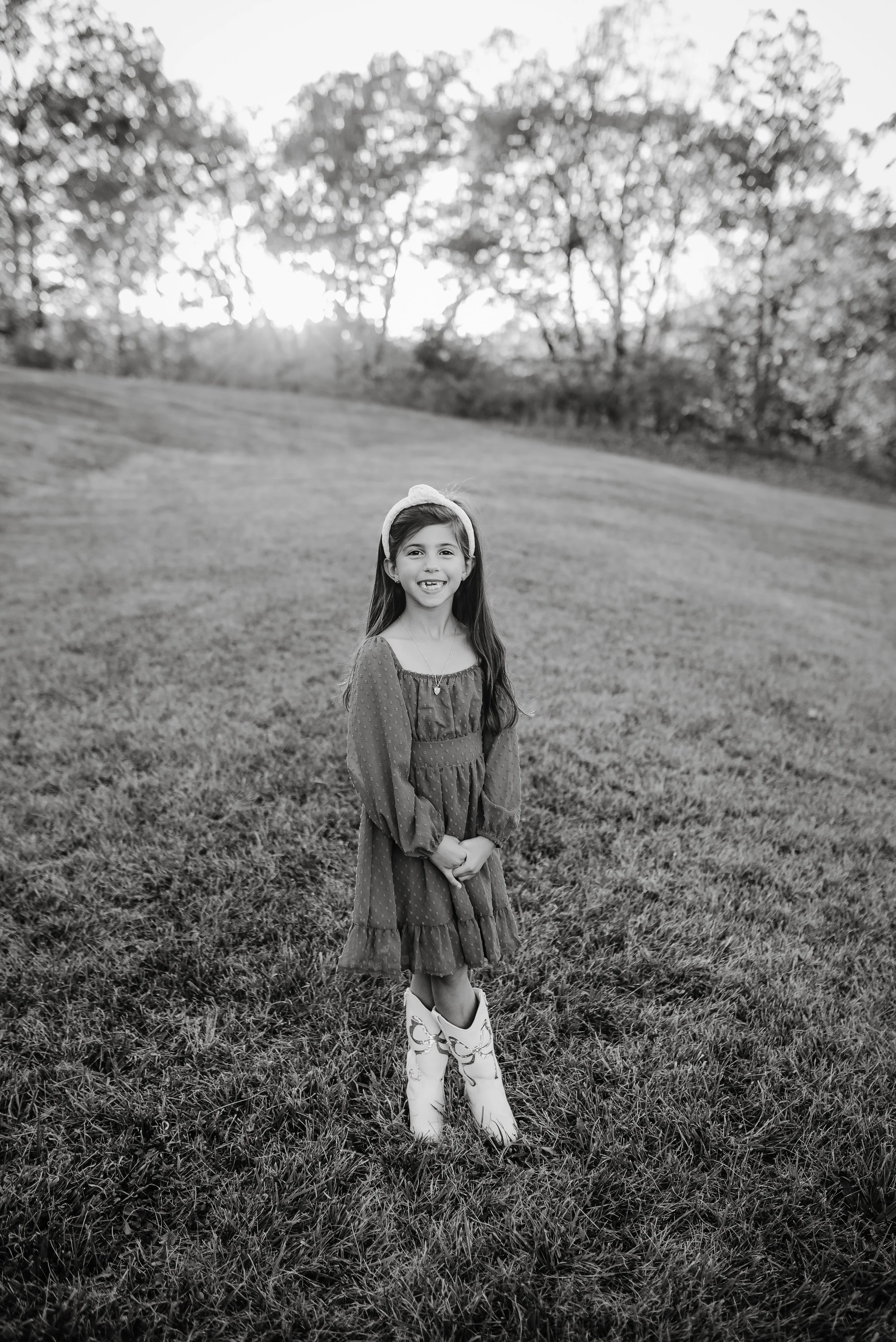 A young girl standing outdoors on a grassy field, smiling with her hands clasped in front of her, wearing a dress, headband, and boots, with trees and sunlight in the background.