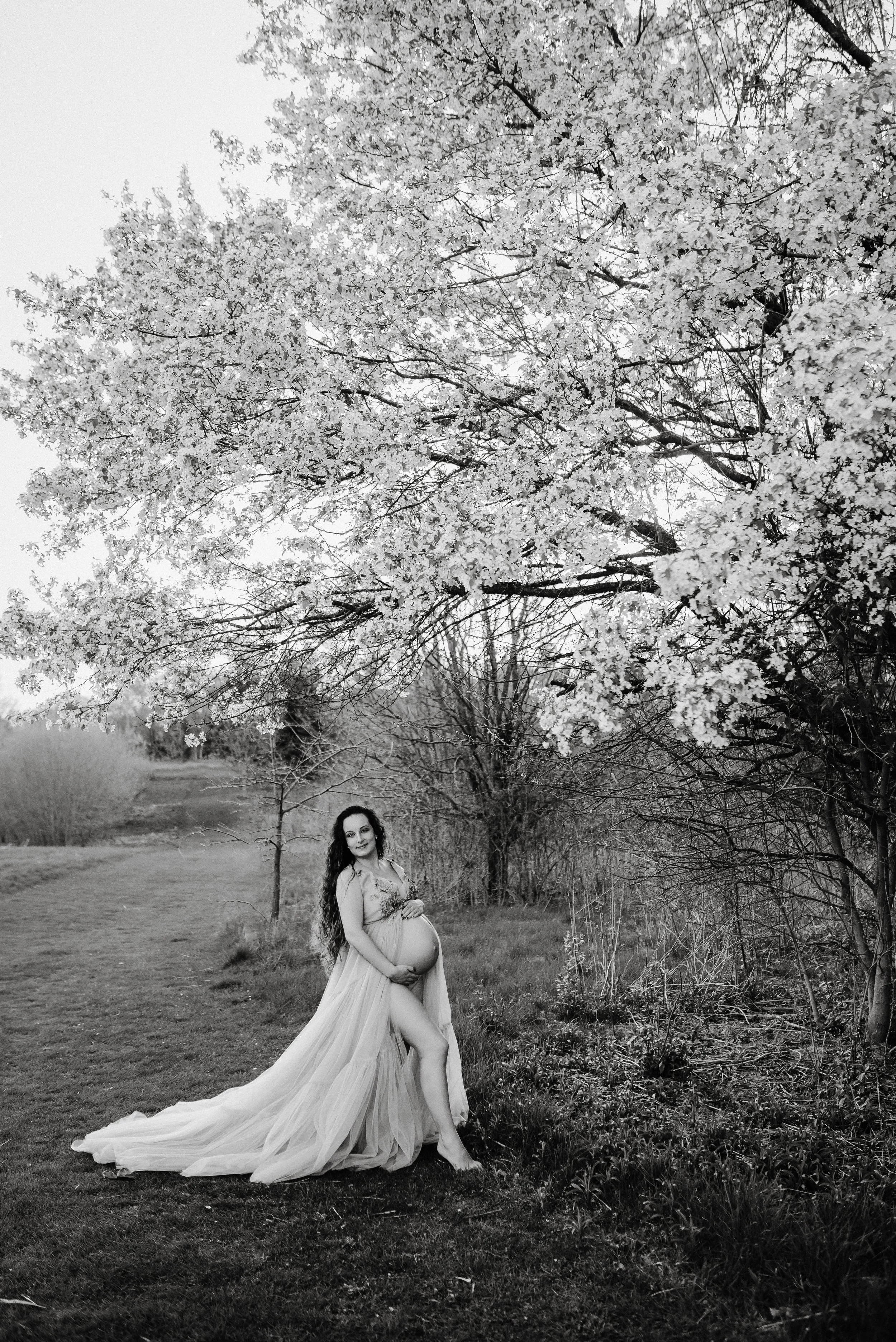 A pregnant woman in a flowing dress stands outdoors near trees with blooming flowers. The photo is in black and white.