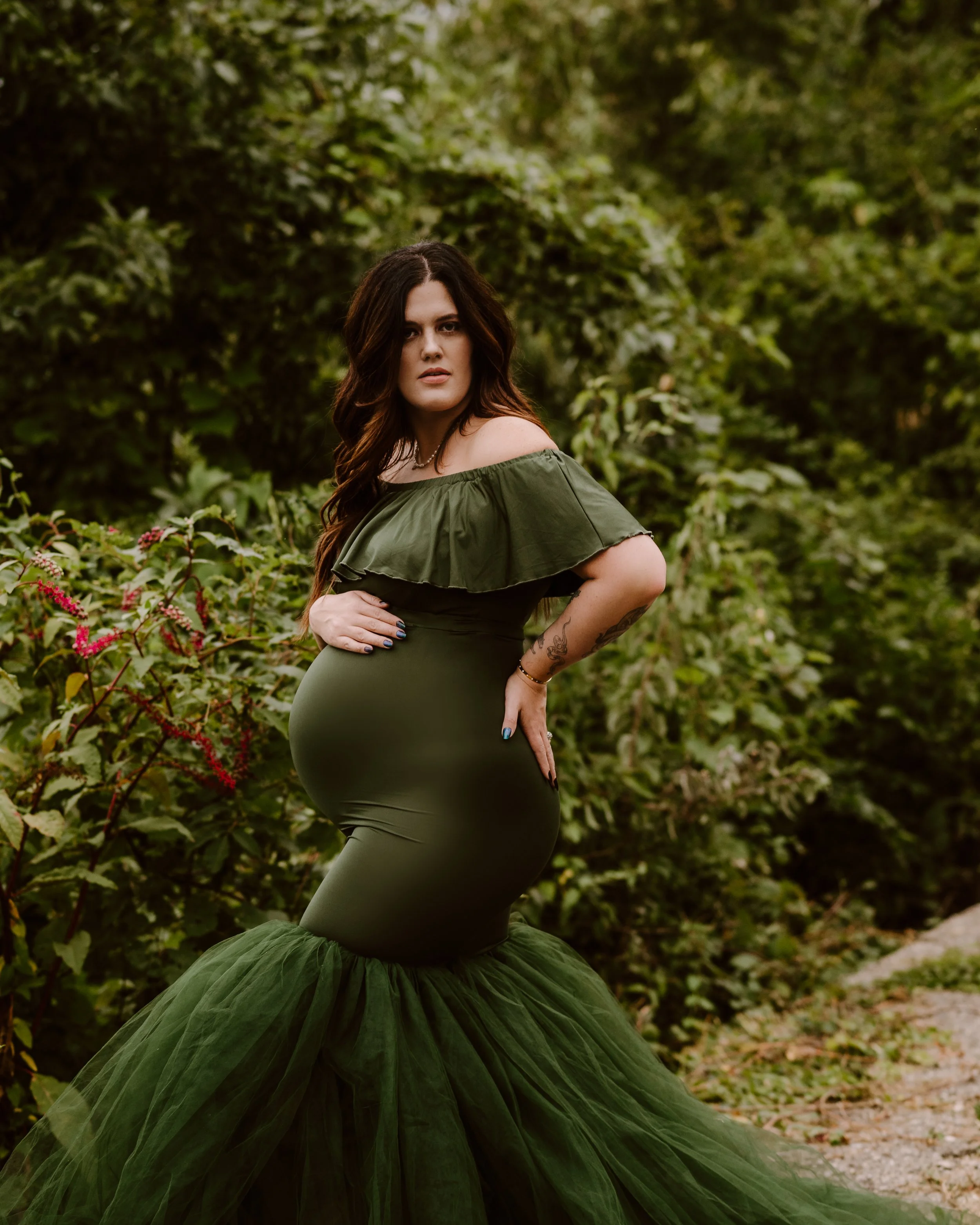 A pregnant woman with long dark hair posing outdoors in a green dress with ruffled off-shoulder top and a tulle skirt, surrounded by lush green foliage.