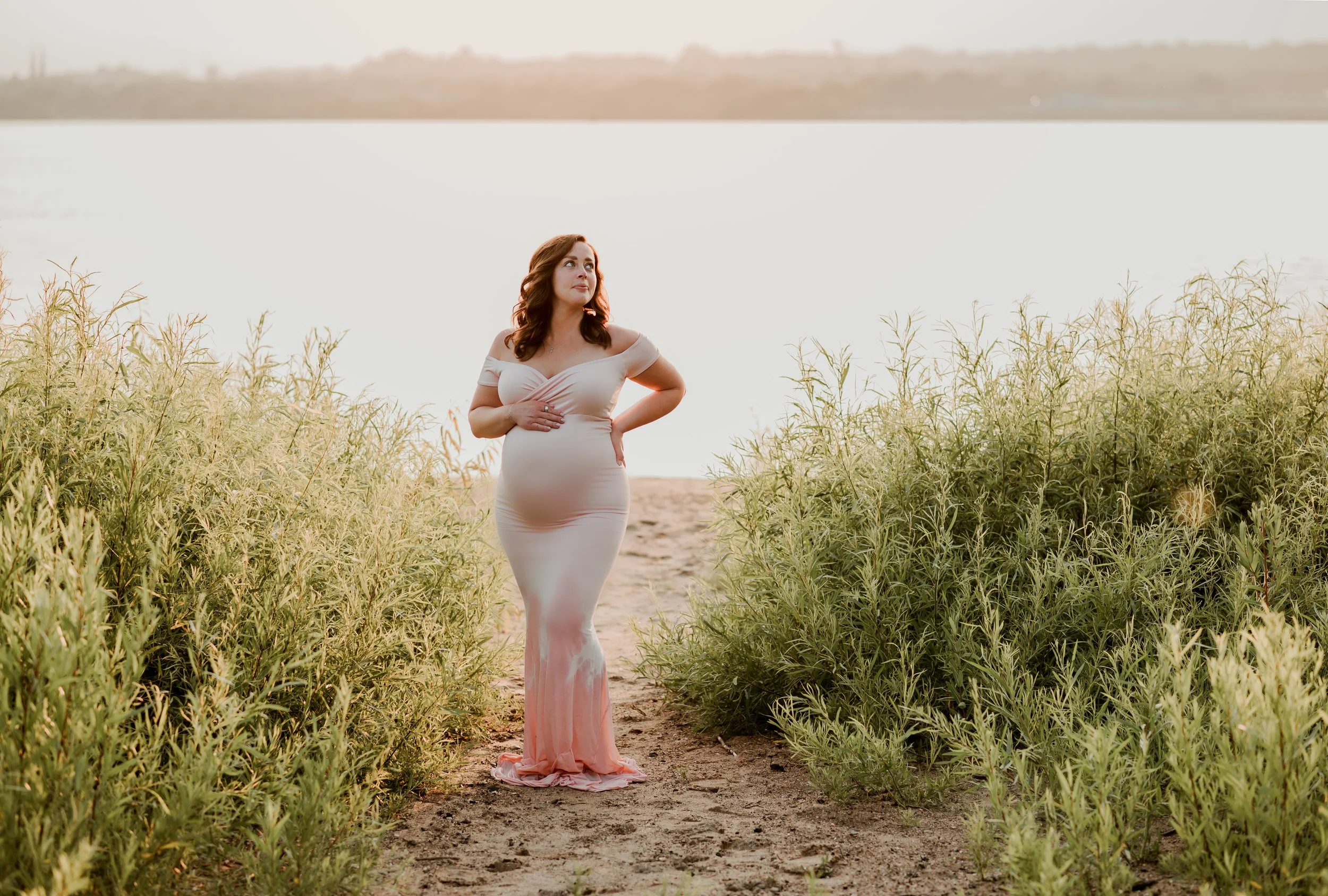 Pregnant woman in a white off-the-shoulder dress standing on a dirt path between green bushes near a body of water during sunset.