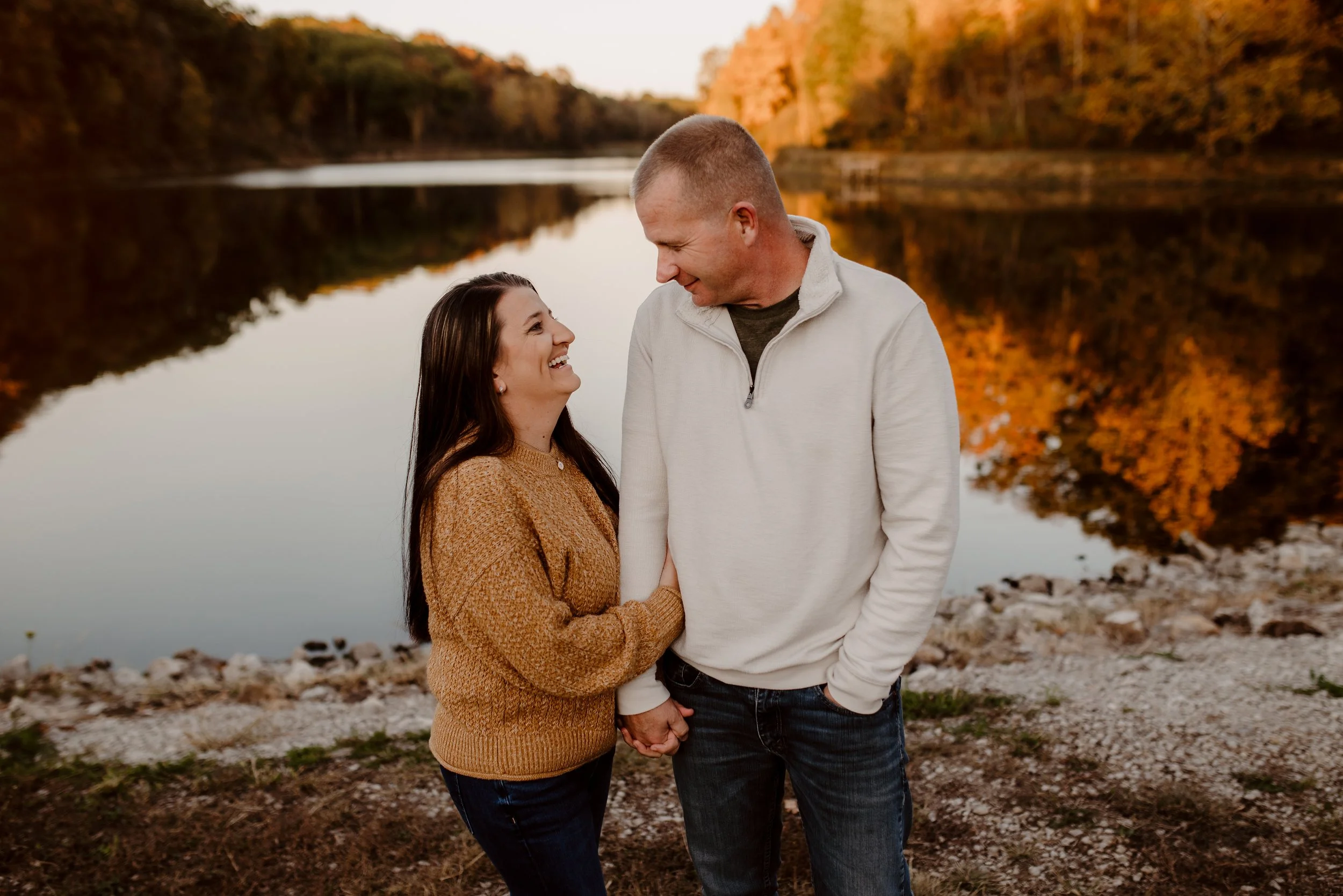 A couple holding hands and smiling at each other by a lake during autumn, with trees with orange and yellow leaves reflected in the water.