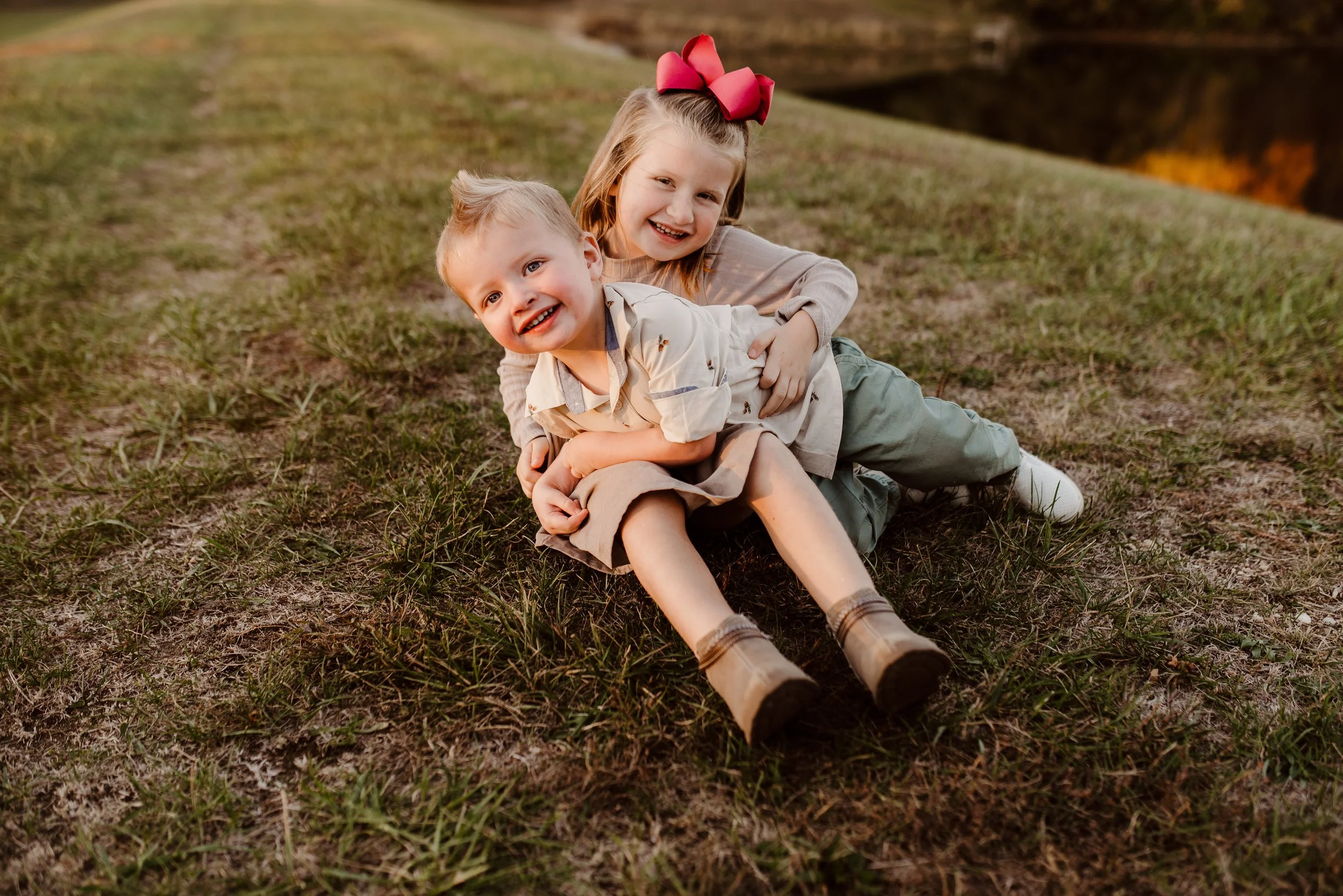 Two young children, a boy and a girl, smiling and playing on grass outdoors during sunset, with the girl sitting behind the boy and hugging him.