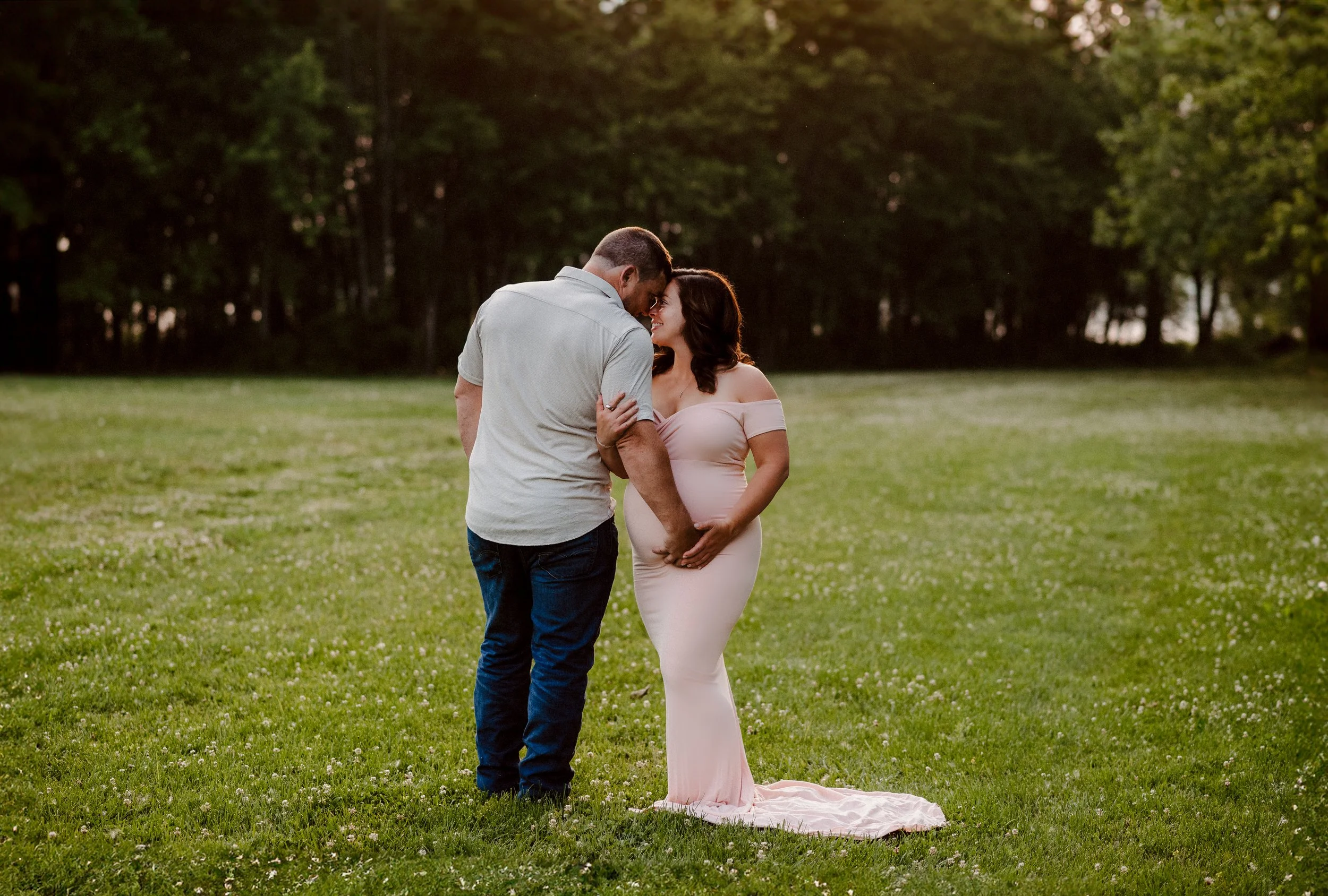 Pregnant woman in a pink dress holding her belly, standing close to a man in a light shirt and jeans, with foreheads touching in a grassy park.
