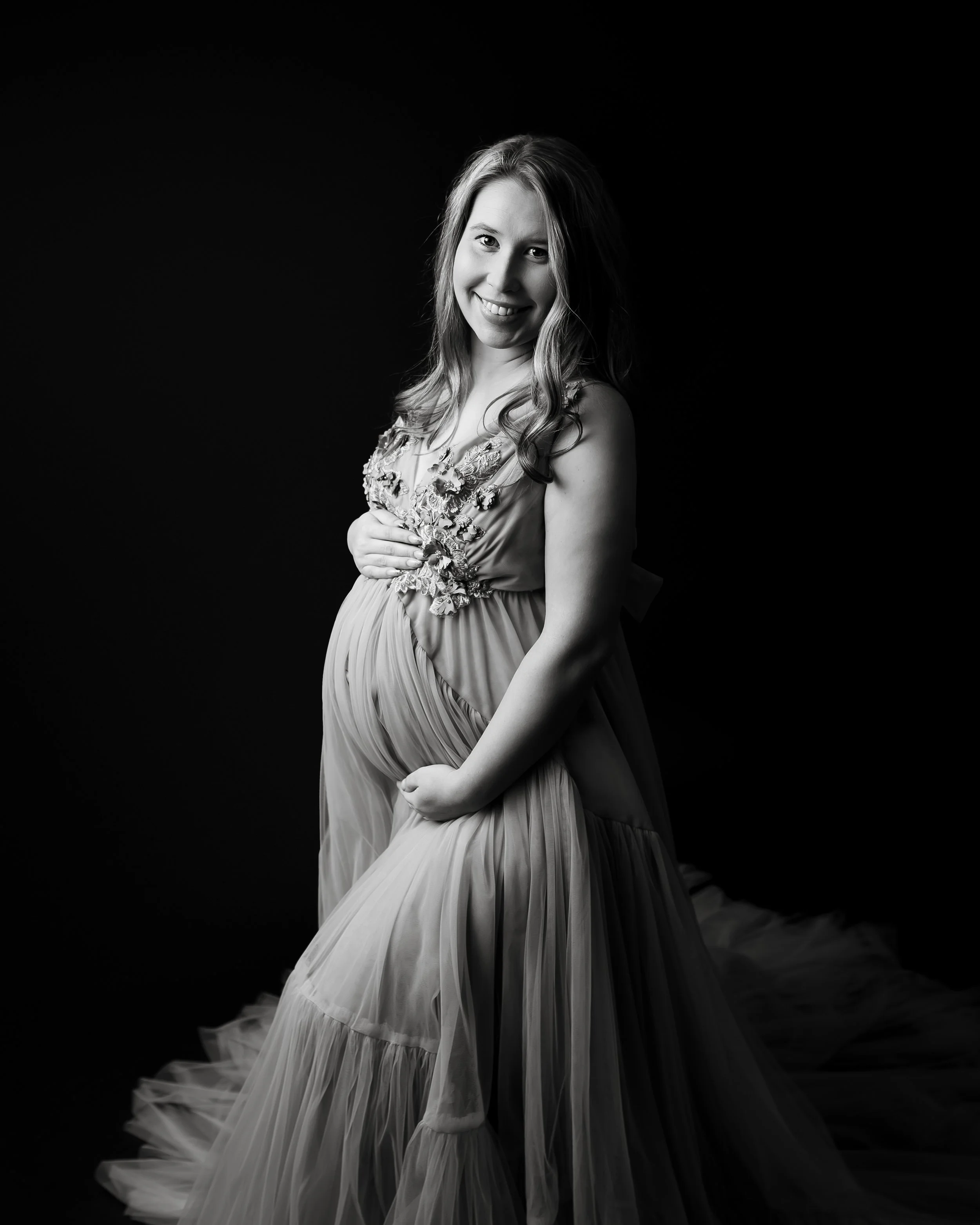 Black and white portrait of a pregnant woman smiling, wearing a flowing gown with floral embellishments, standing against a dark background.