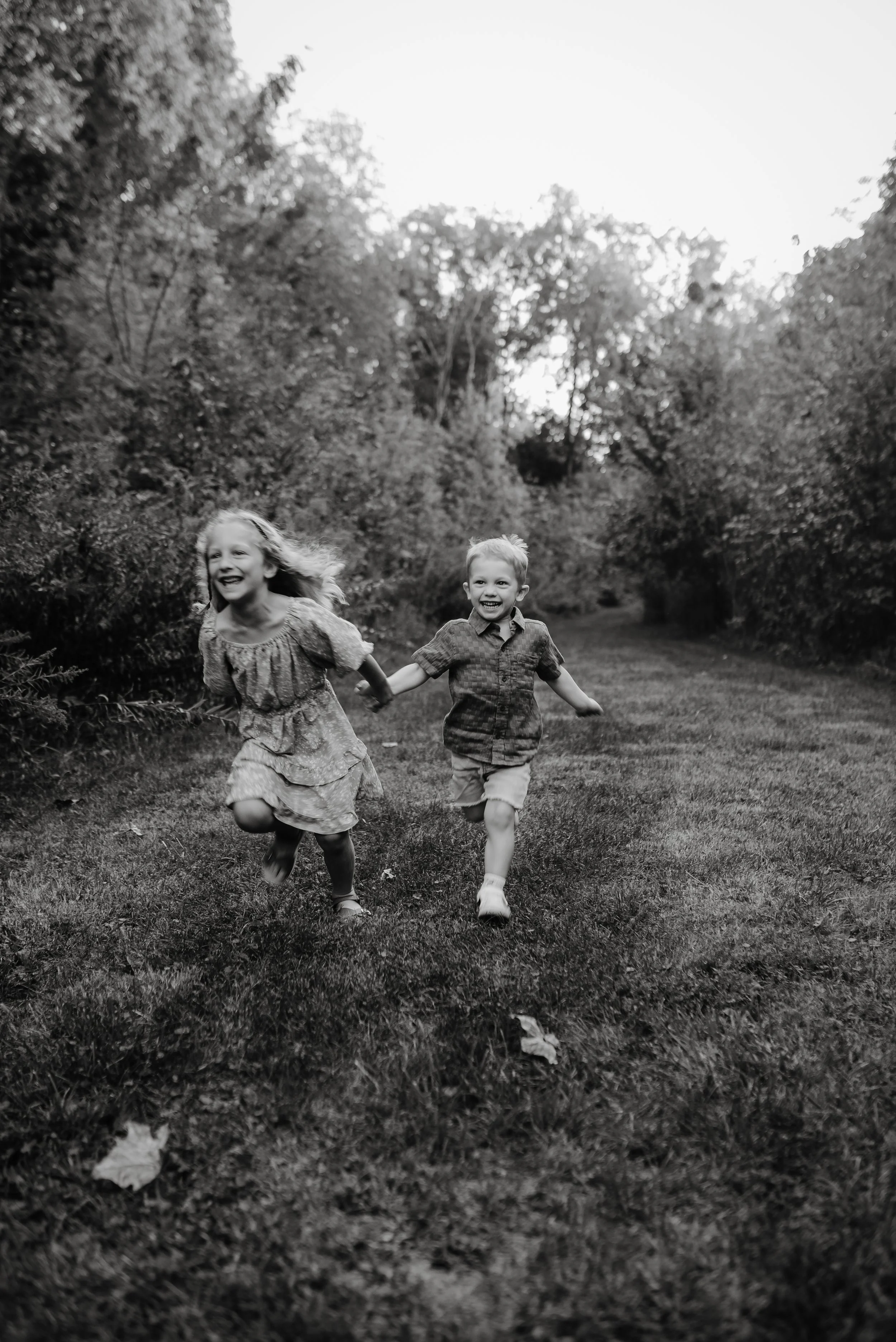 Two children, a girl and a boy, happily running and holding hands along a grassy path surrounded by trees, in black and white.