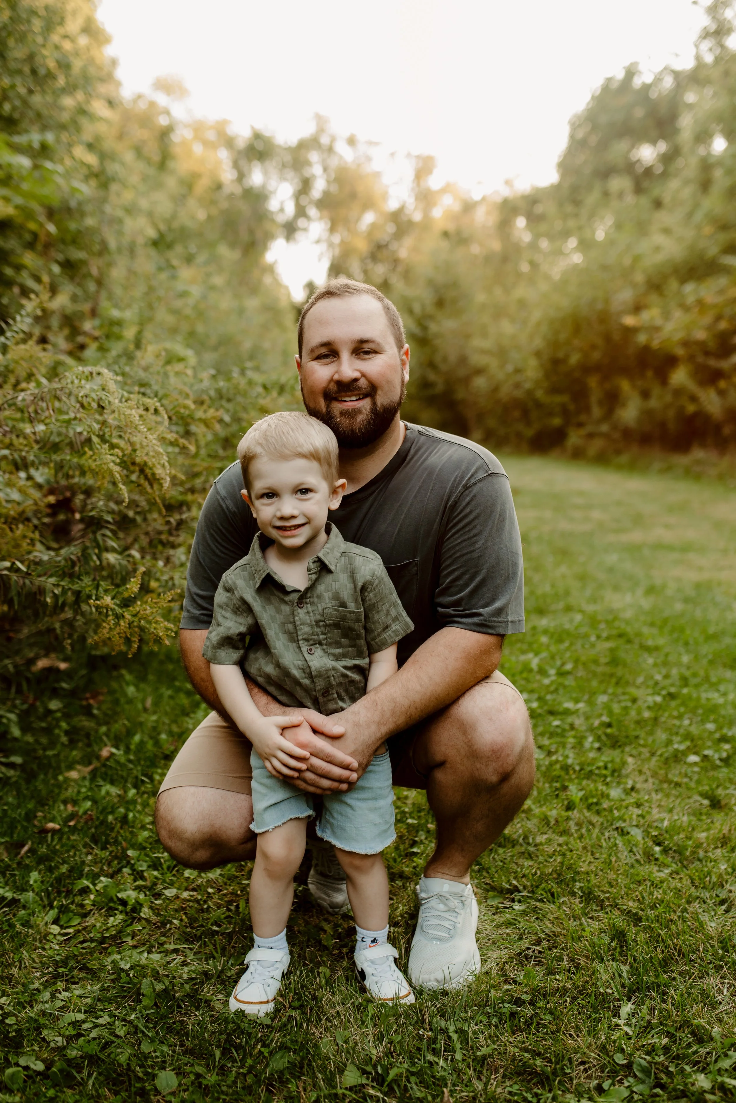 A man and a young boy outdoors on a grassy trail, smiling at the camera. The man is squatting behind the boy, holding him by the waist. The boy is wearing a green shirt, shorts, and sneakers, and the man is wearing a dark t-shirt, shorts, and sneaker