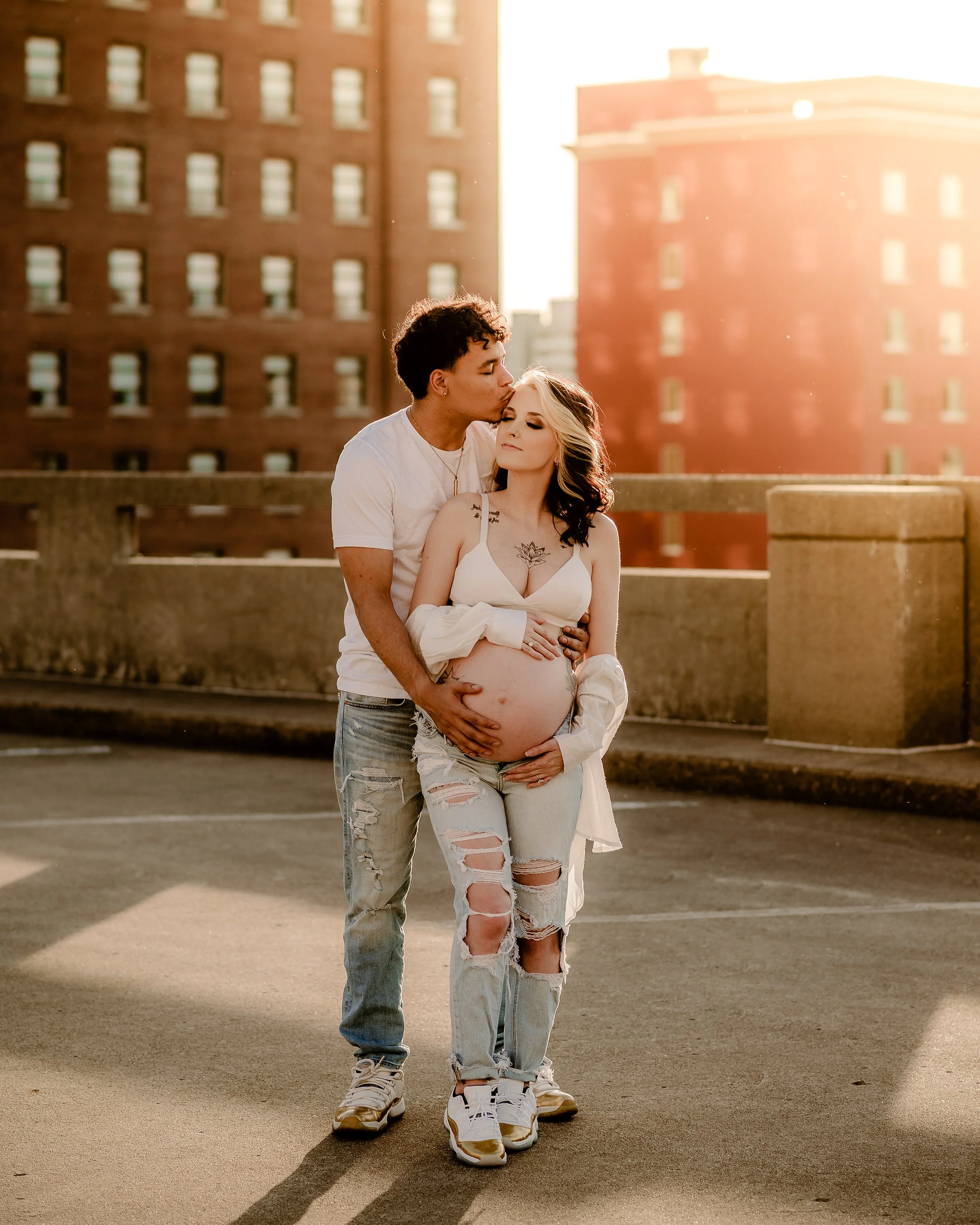 A pregnant woman with dark hair in a white crop top and ripped jeans stands with a man on a rooftop parking lot, sunlight shining behind them. The man, with curly hair, kisses her on the forehead, and they are holding her belly.