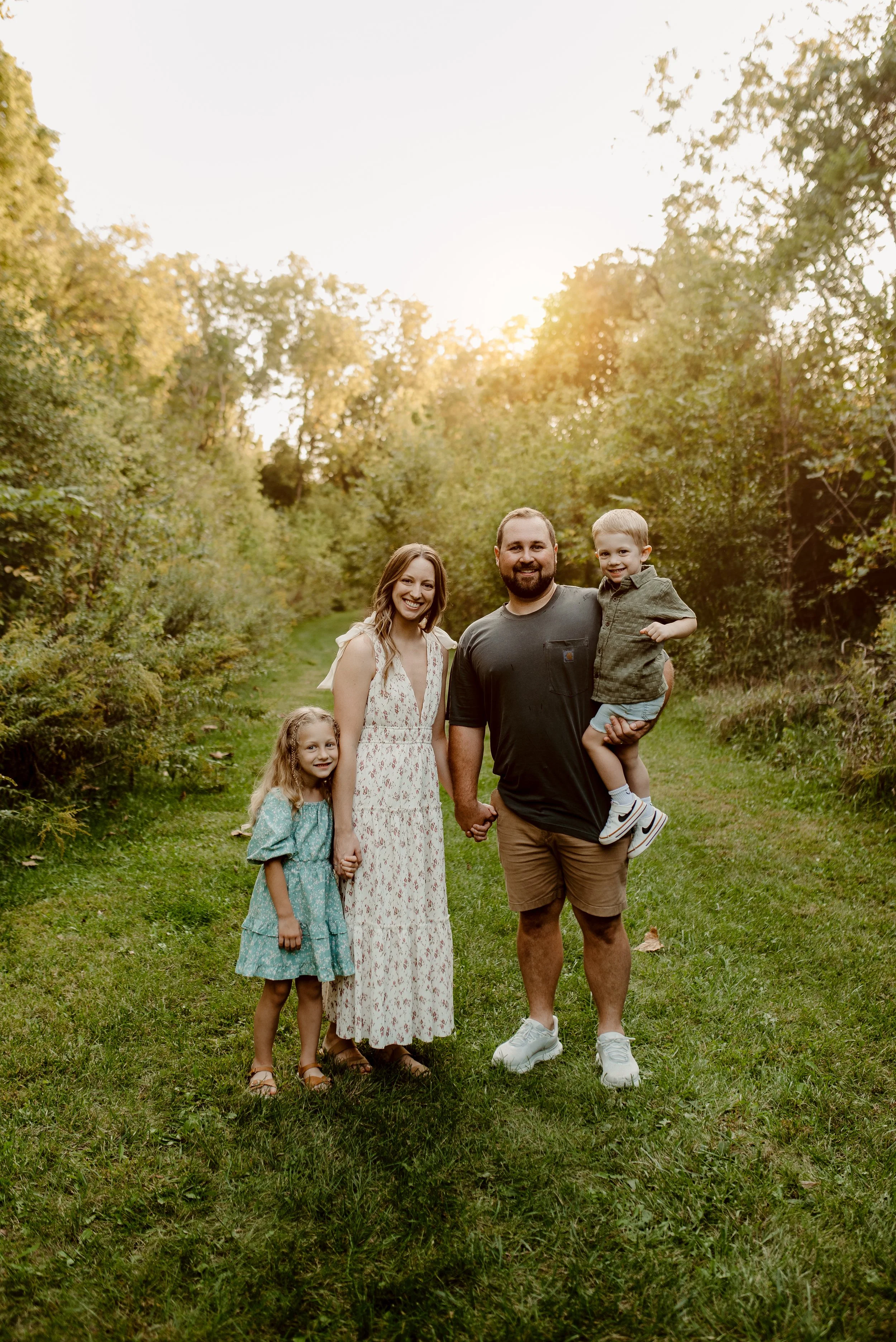 A family of four holds hands on a grassy path in a wooded area during sunset, smiling at the camera.
