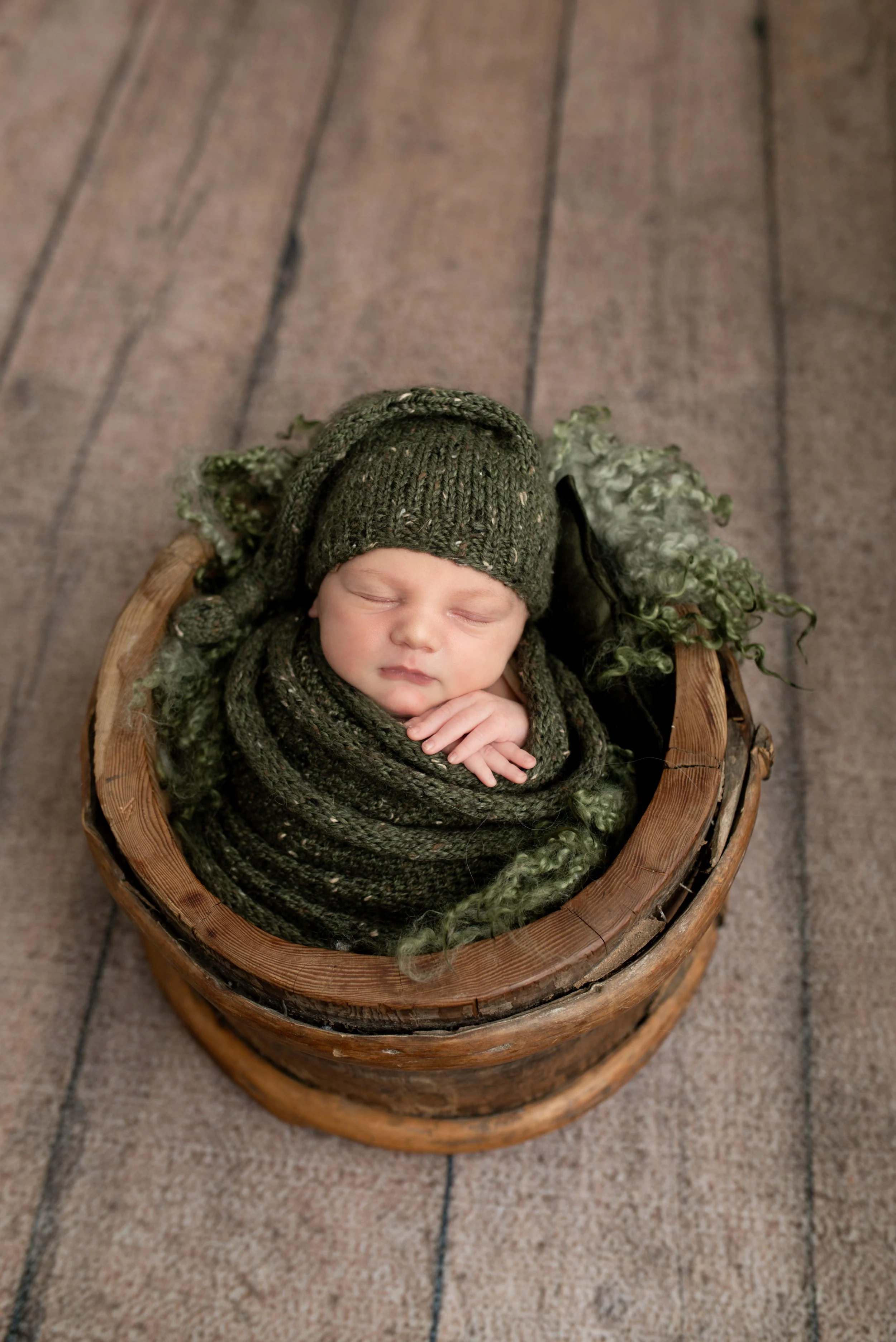 A sleeping baby wrapped in a dark green knitted blanket and wearing a matching hat, lying in a rustic wooden bowl on a wooden floor.