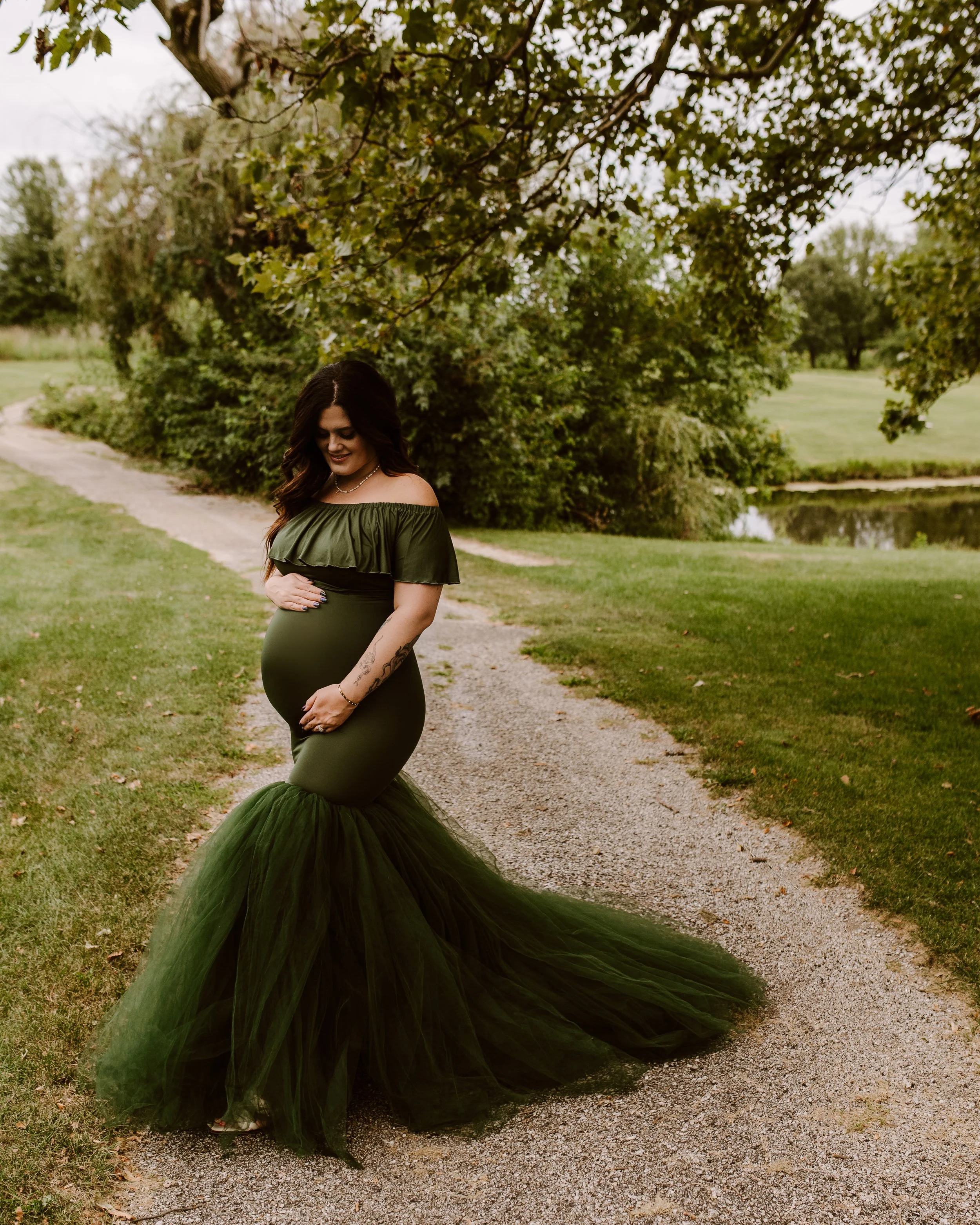 A pregnant woman dressed in a dark green off-shoulder gown with a ruffled top and a tulle train, standing on a gravel path in a park with greenery and a pond in the background, smiling while holding her belly.