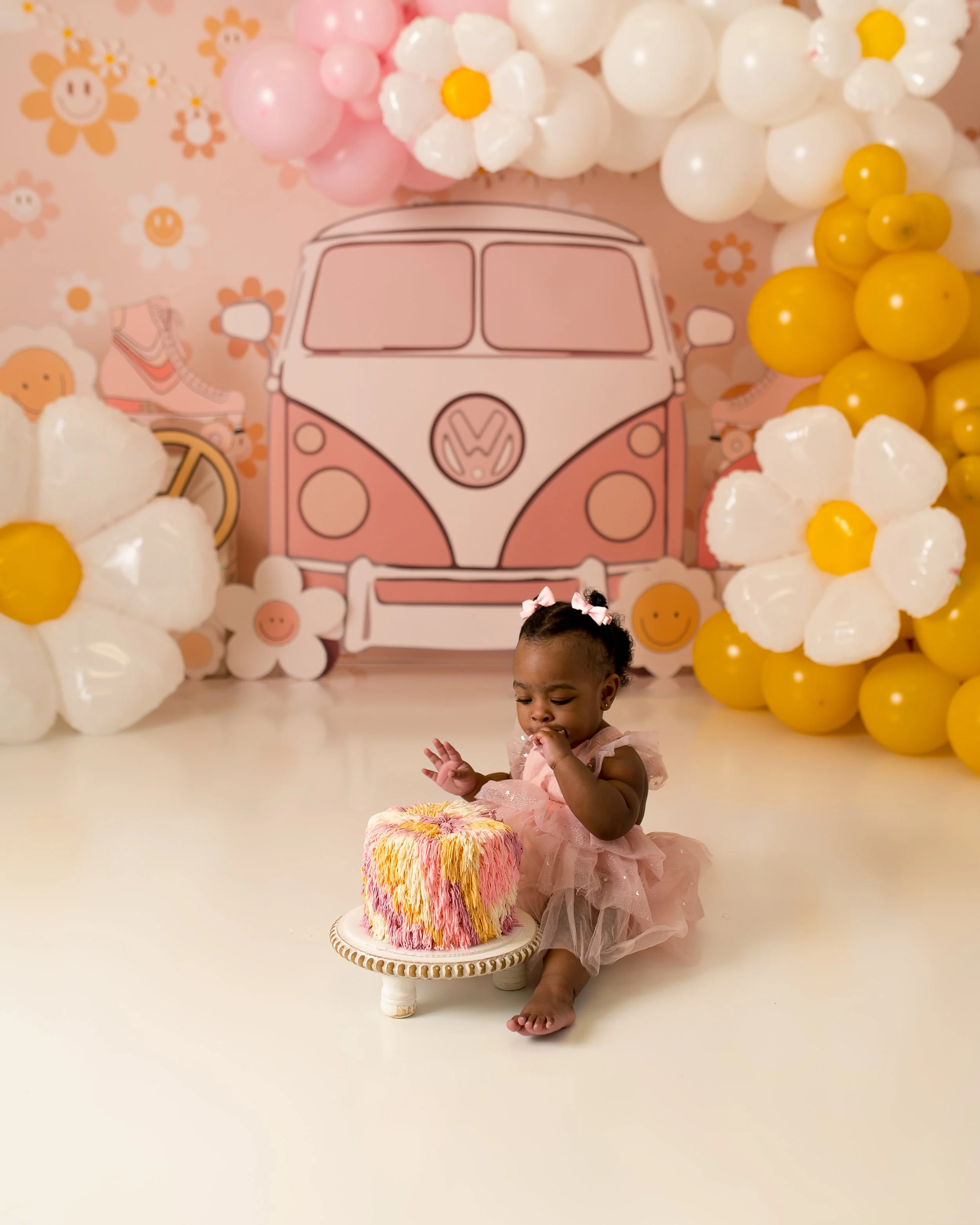 A young girl in a pink dress sitting on the floor, holding a pink and yellow birthday cake, with colorful balloon arrangements and a cartoon van backdrop in the background.