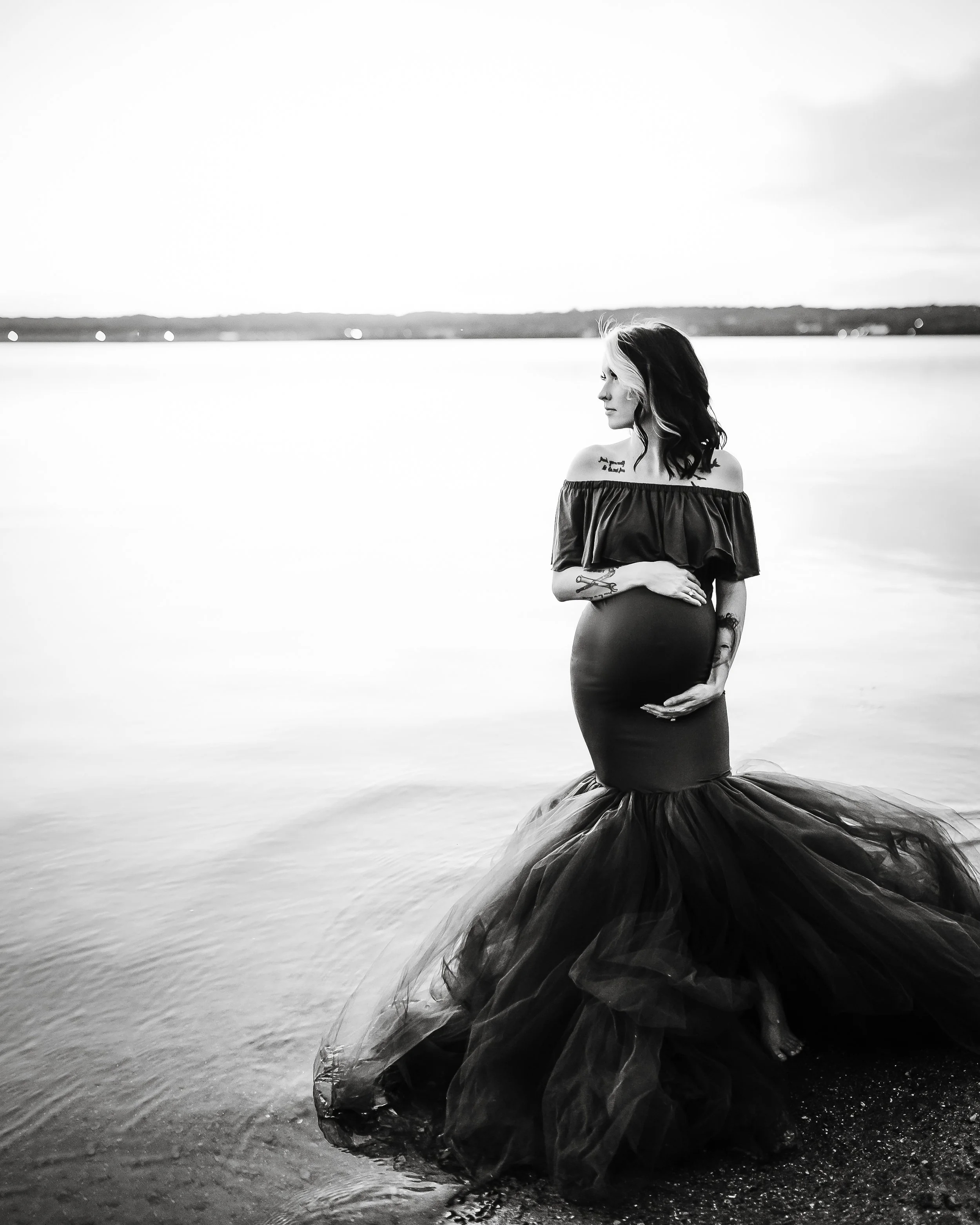 A woman in a black off-the-shoulder gown standing at the edge of a body of water, looking to the side, with her hand on her stomach, in a black-and-white photo.