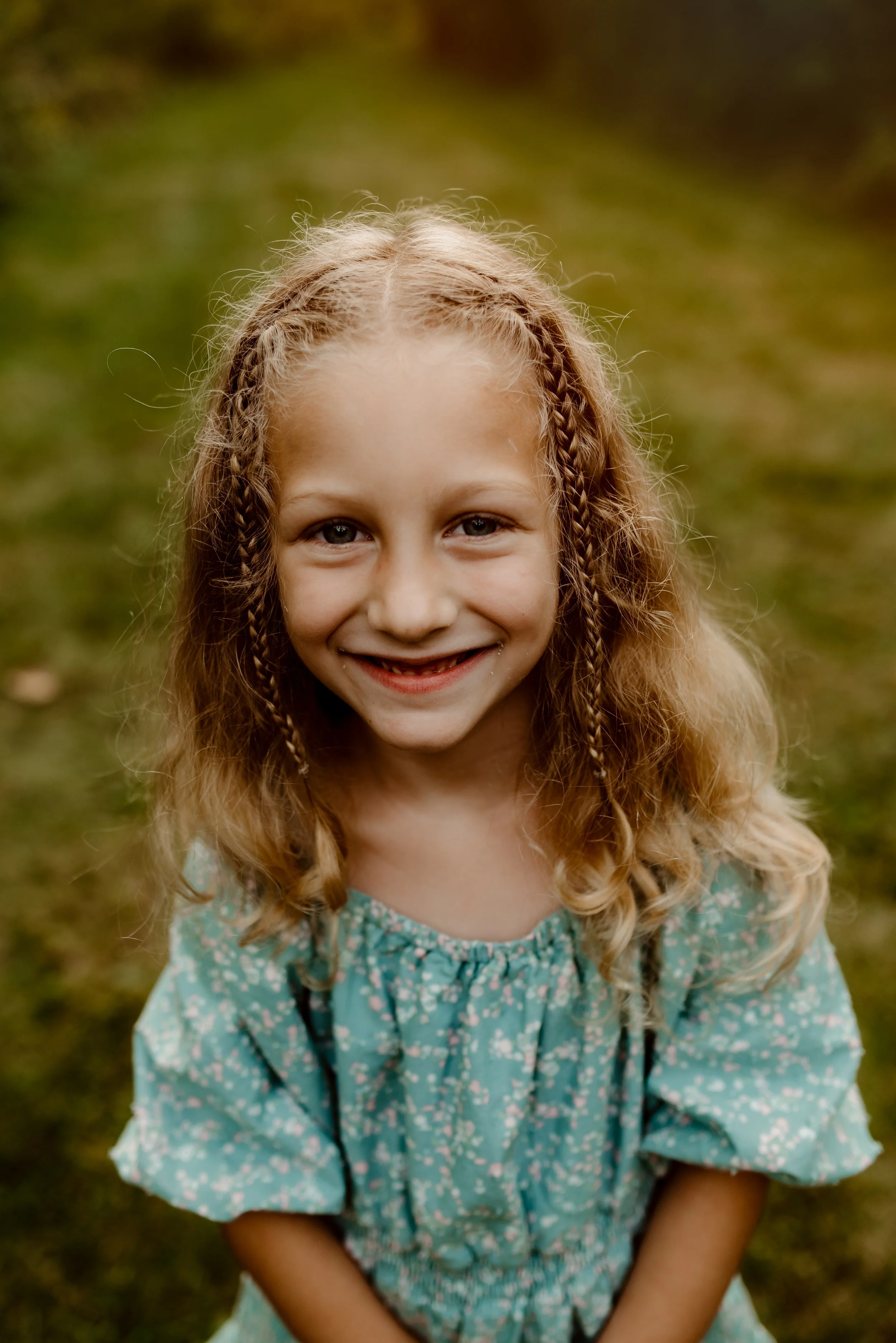 A young girl with curly blonde hair and a big smile, wearing a blue floral dress, standing outdoors on a grassy path.