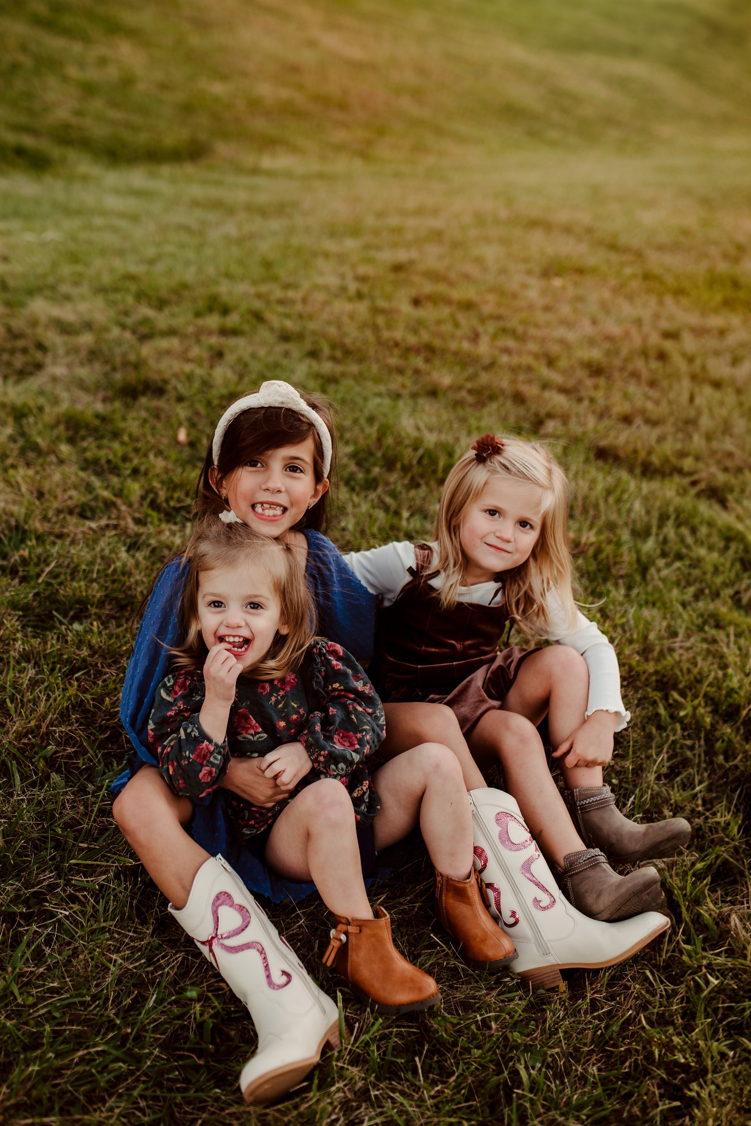 Three young girls sitting on the grass, smiling and enjoying the outdoors during golden hour with warm lighting.