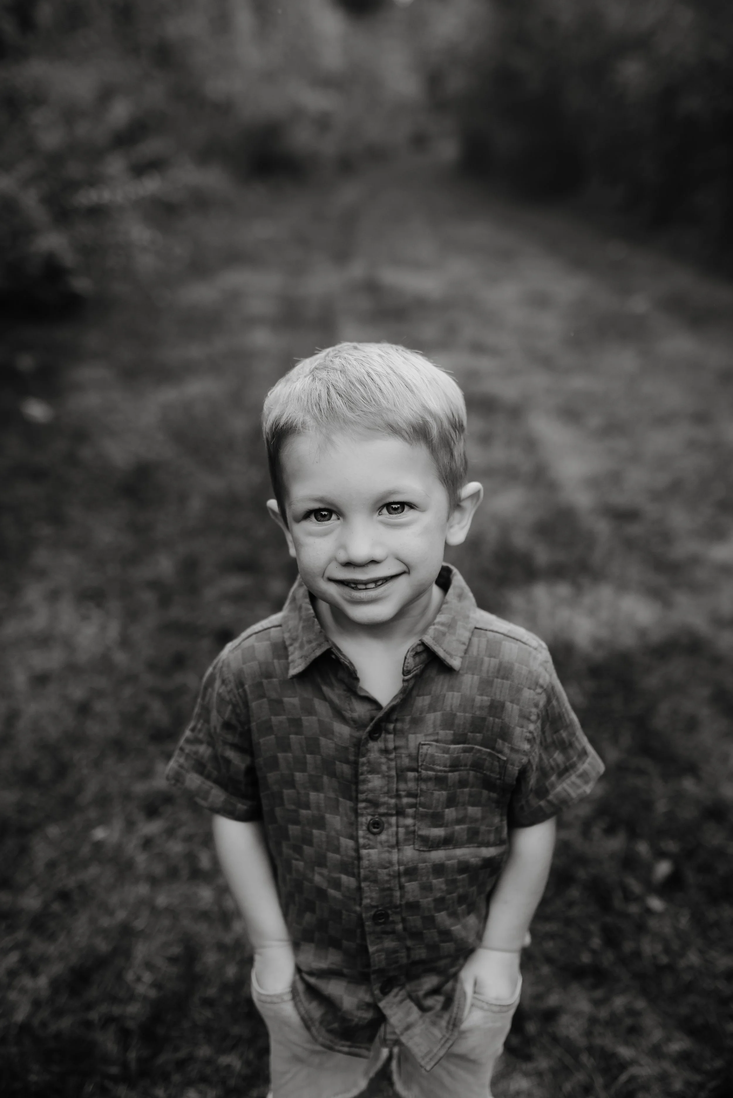 A young boy smiling outdoors in a park, facing the camera with hands in his pockets, in black and white.