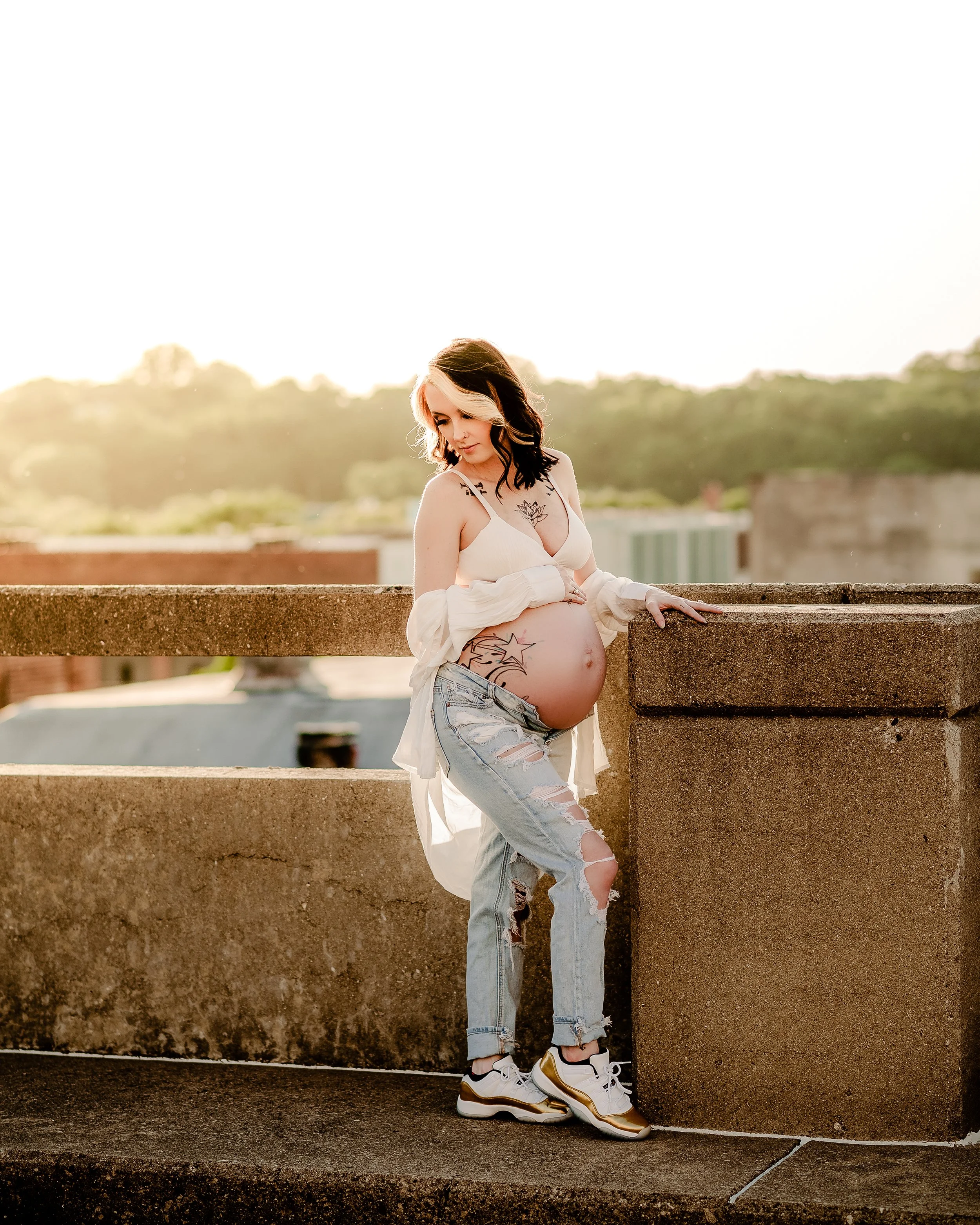 Pregnant woman with tattoos and blonde and dark hair, wearing a white top, ripped jeans, and sneakers, standing on a rooftop at sunset