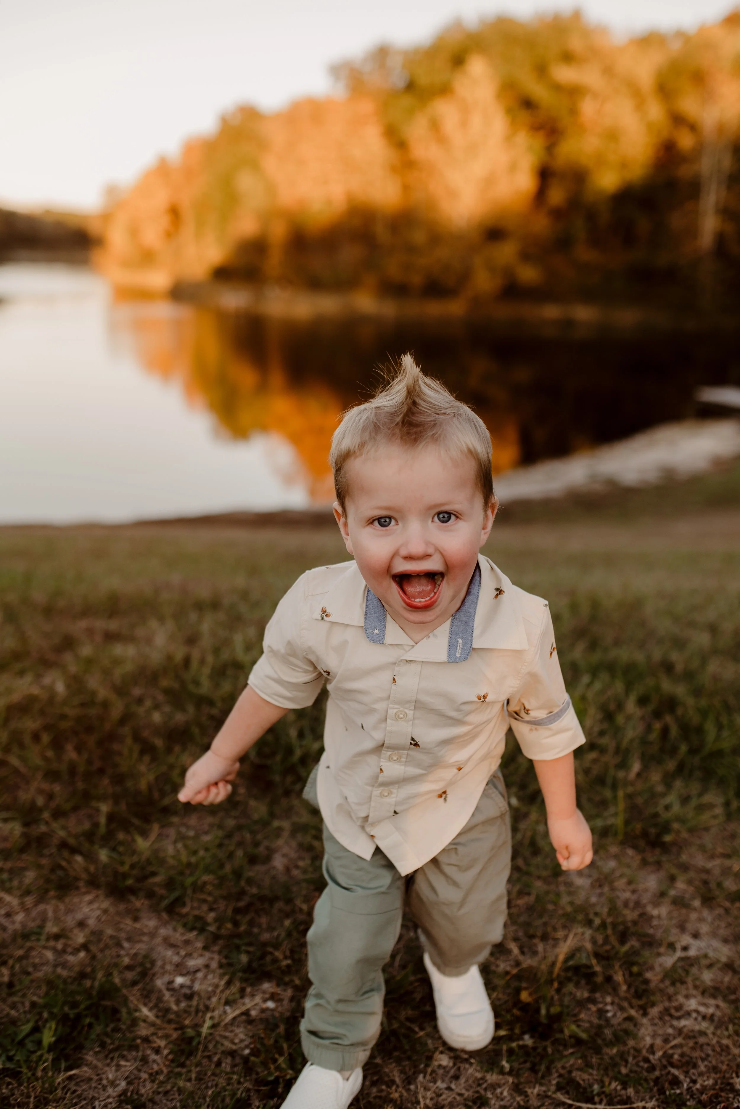 A young boy with a mohawk hairstyle, wearing a beige shirt with small animal prints, khaki pants, and white shoes, is running and smiling excitedly outdoors near a body of water with autumn trees in the background.