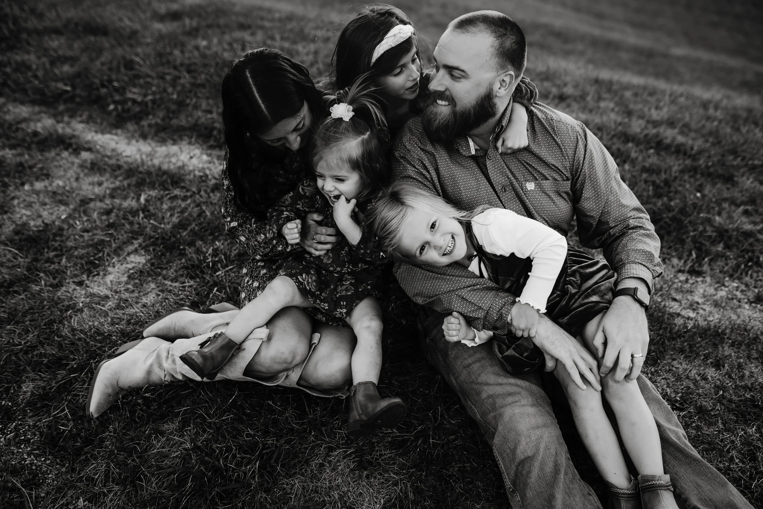Black and white photo of a family of six sitting on the grass, smiling and laughing together. The family includes two adult men and women, and four young girls. They are closely huddled, showing affection and joy.