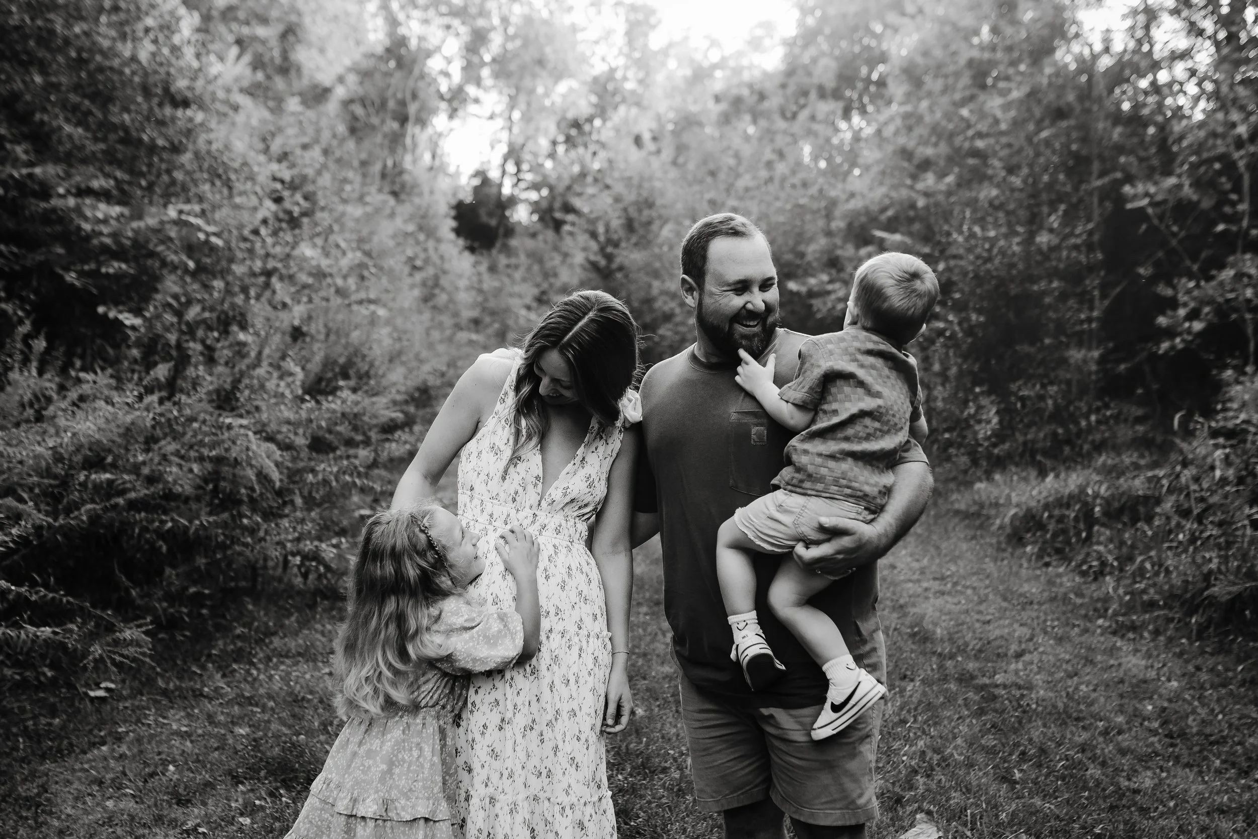 A family of four, including a man, woman, and two young children, walking together outdoors on a wooded path, enjoying a joyful moment.