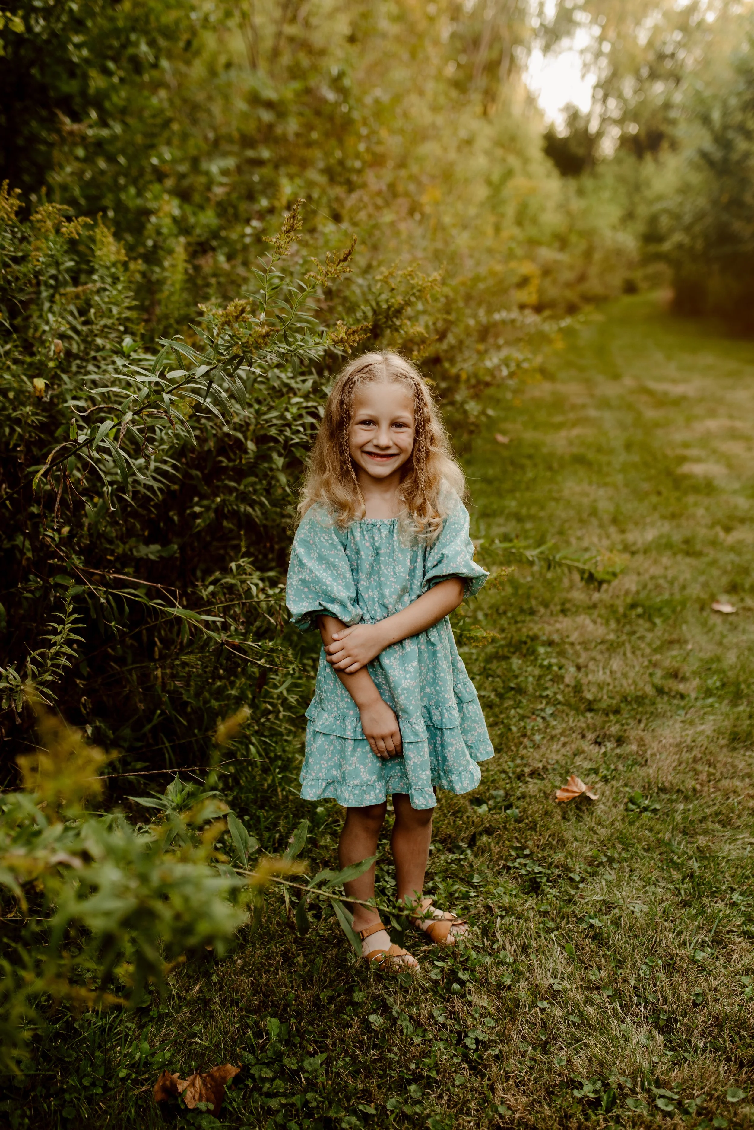 A young girl with curly blonde hair in a blue, floral dress standing on a grassy path surrounded by green bushes and trees, smiling at the camera during daytime.