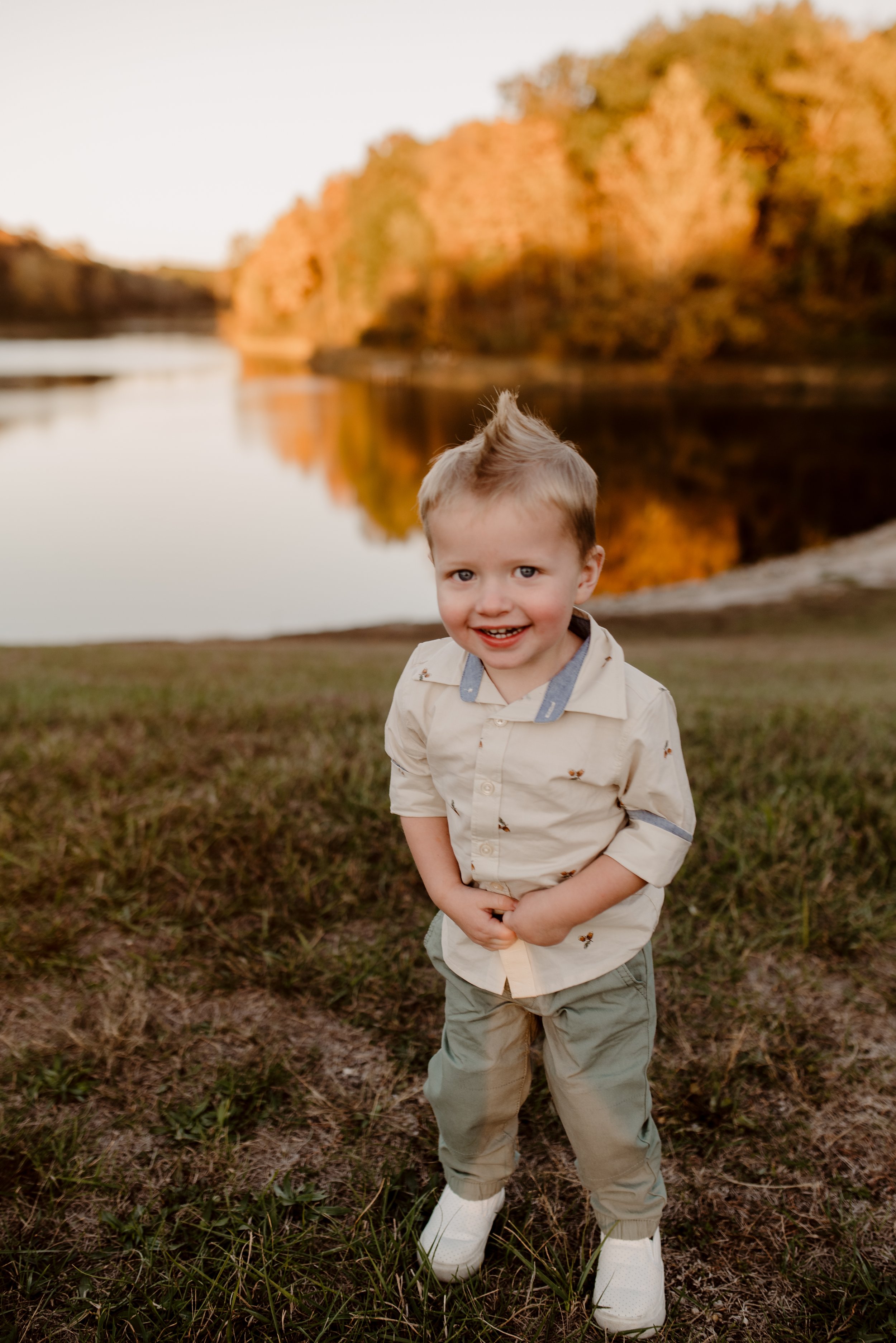 A young boy with blonde hair smiling outdoors near a river during fall with orange and yellow trees.