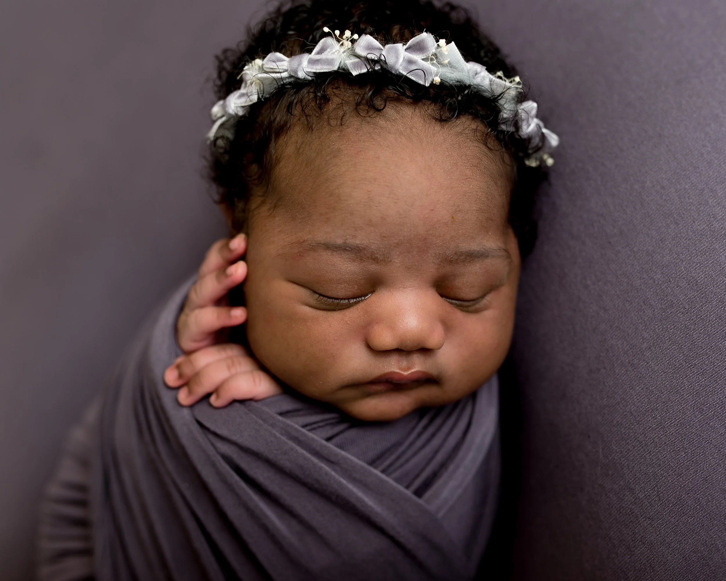 A close-up of a sleeping baby girl with a black curly hair, wearing a white floral headband, wrapped in a dark gray blanket, lying against a gray background.