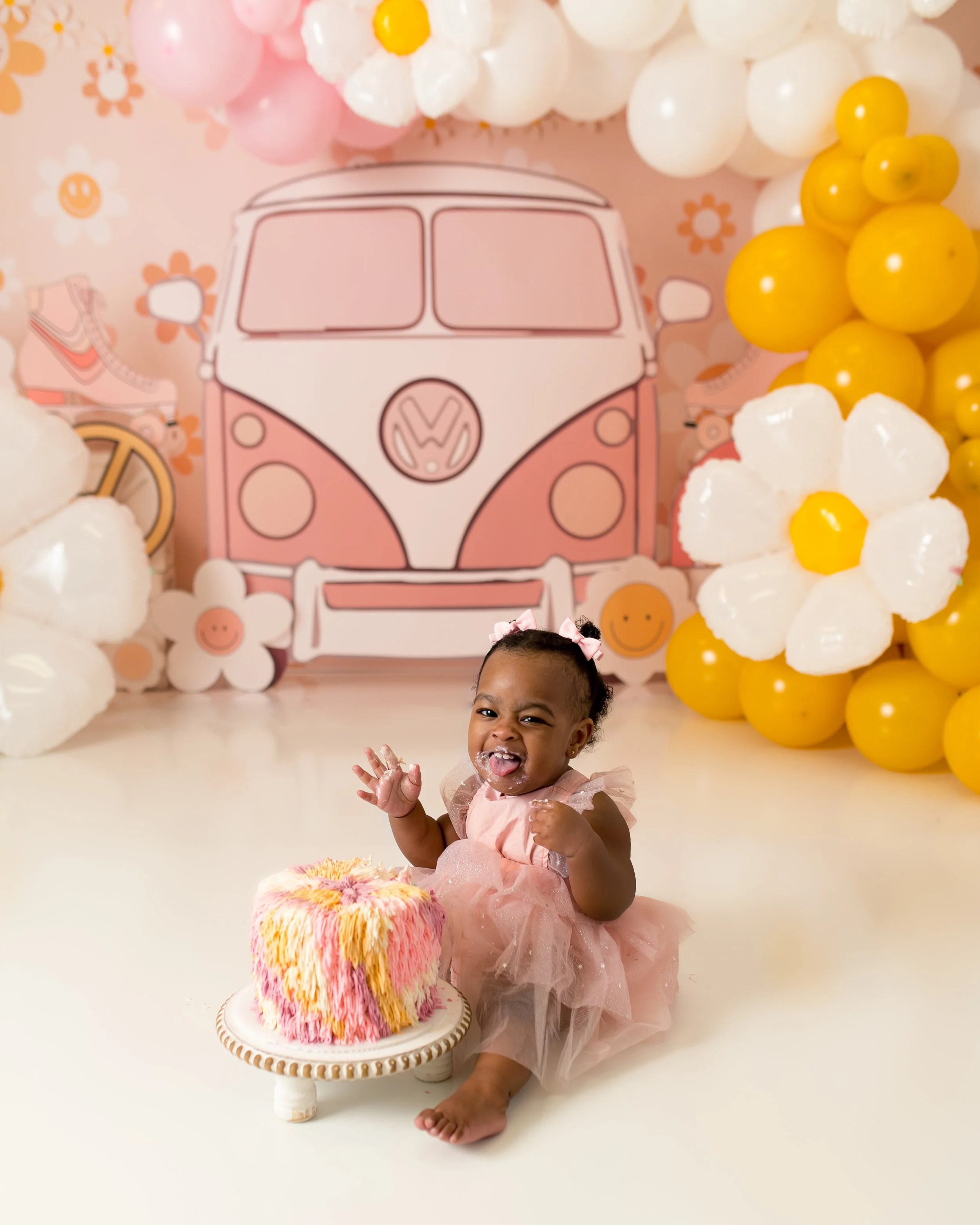 A smiling young girl in a pink dress sitting on the floor with a colorful birthday cake in front of her, celebrating her first birthday.