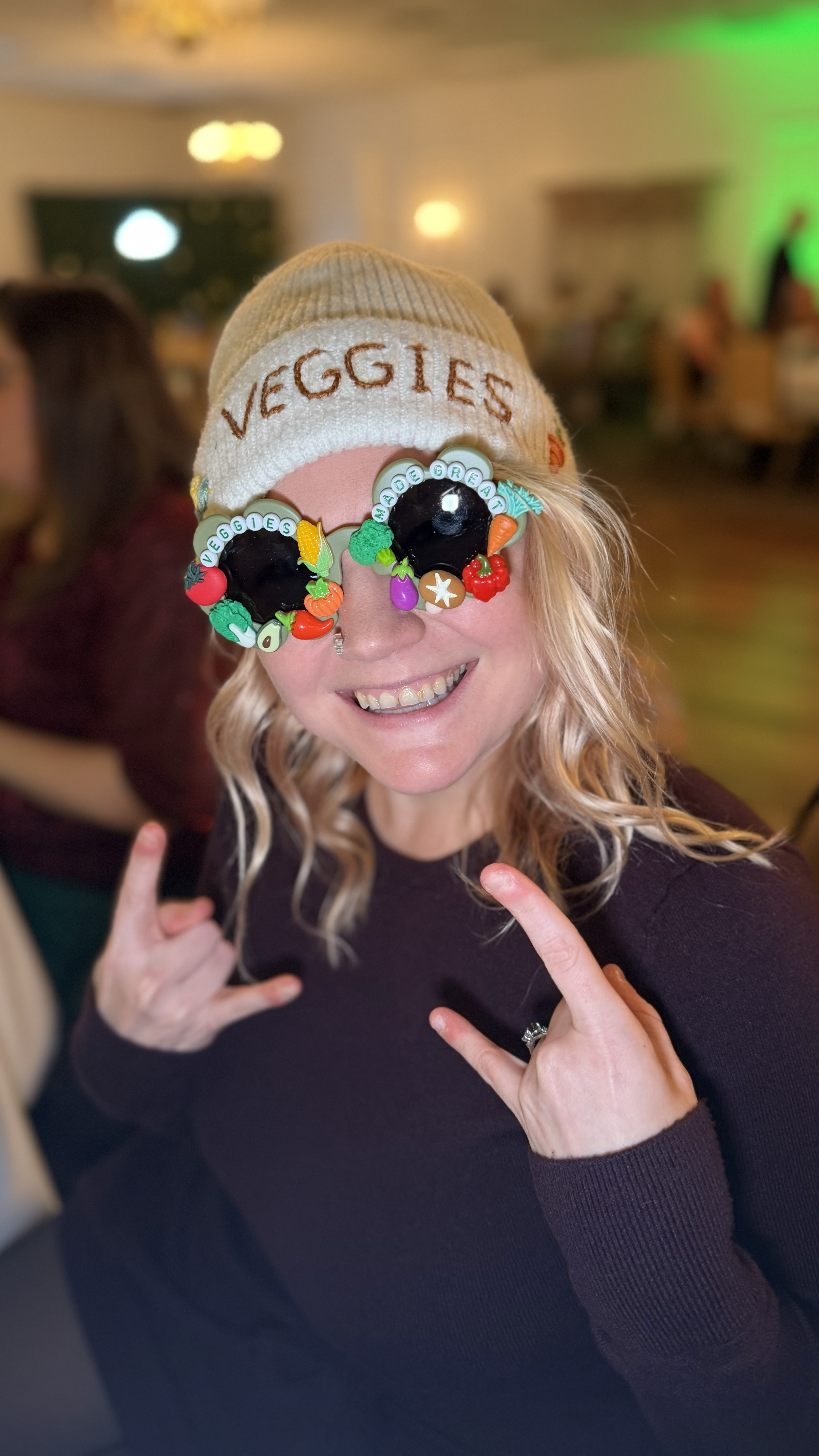 A woman smiling and making rock on hand gestures while wearing colorful veggie-themed sunglasses and a beige beanie with 'VEGGIES' written on it, in a lively indoor setting.