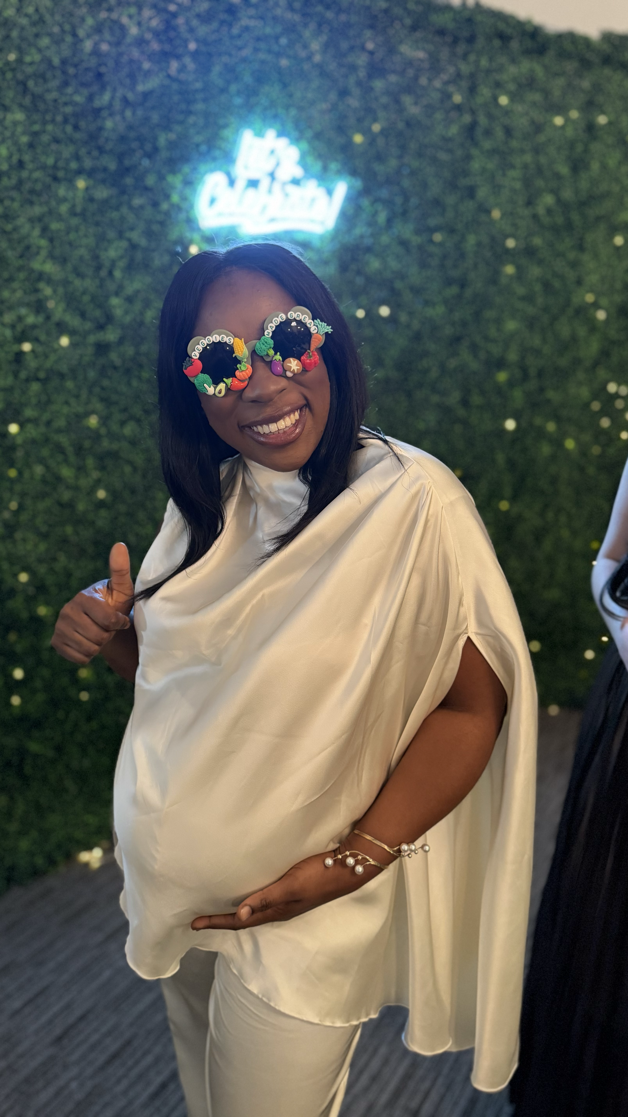 Woman with dark hair wearing silk cream-colored dress and colorful beaded glasses, smiling and giving a thumbs up, standing in front of a green hedge with a neon sign that reads 'Let's Celebrate.