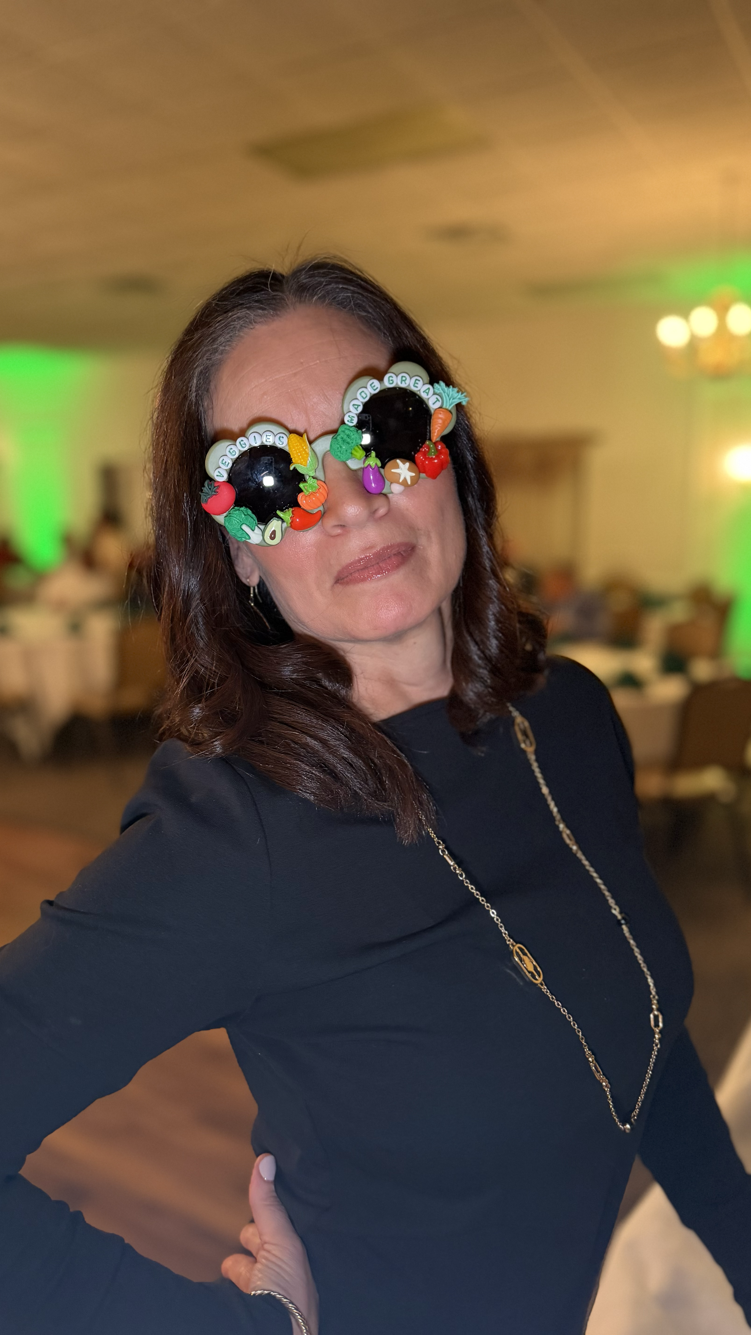 A woman wearing novelty sunglasses with colorful fruit decorations and the words 'VEGGIES MADE GREAT' at an indoor event.