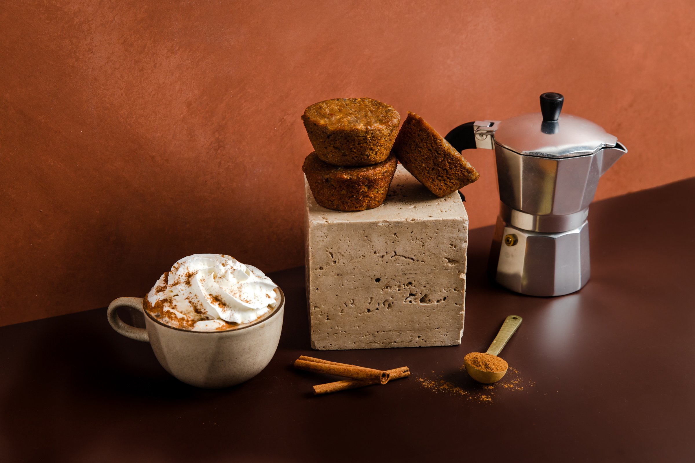 three keto friendly cinnamon roll muffins stacked on a concrete block next to a cup of coffee with whipped cream