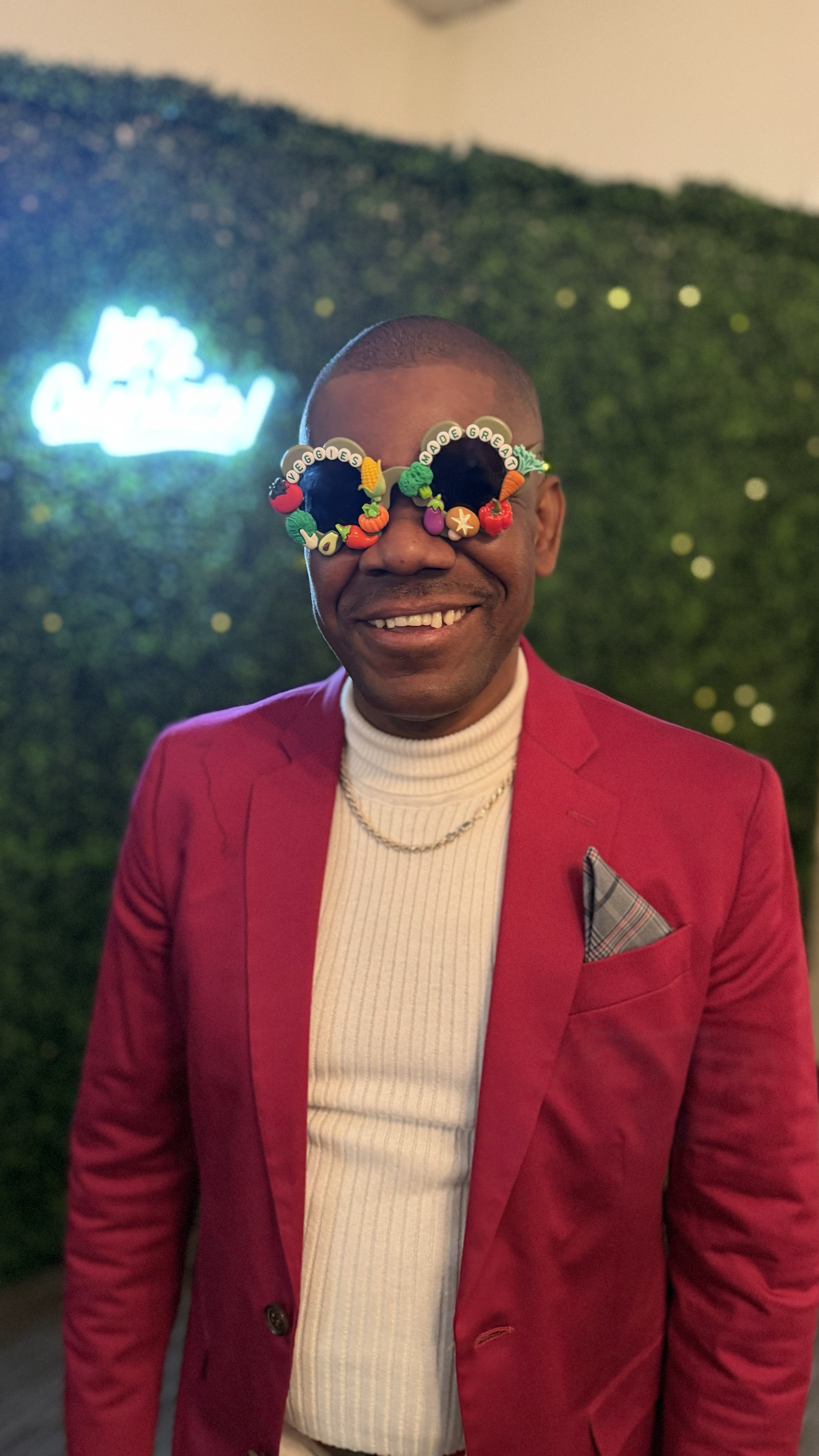 A smiling man wearing sunglasses decorated with small colorful vegetable embellishments, a red blazer, a white turtleneck sweater, and a chain necklace, standing in front of a green, leaf-covered wall with a neon sign in the background.