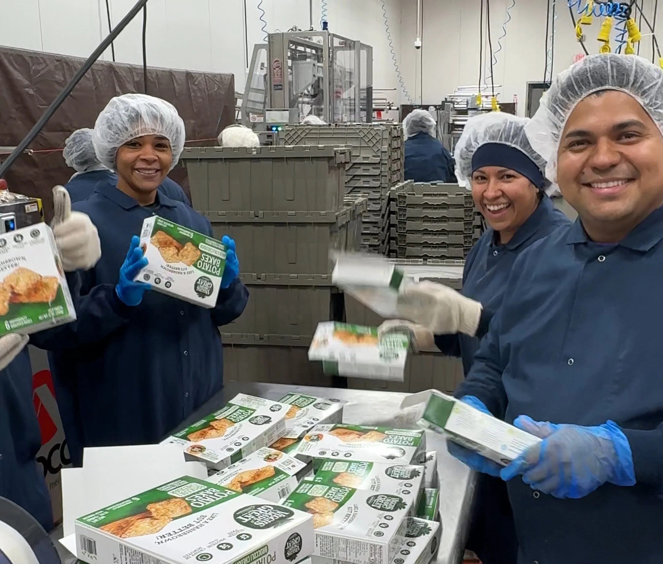 Workers in hairnets and gloves packaging veggies made great products. They are smiling and handling boxes in a factory setting.