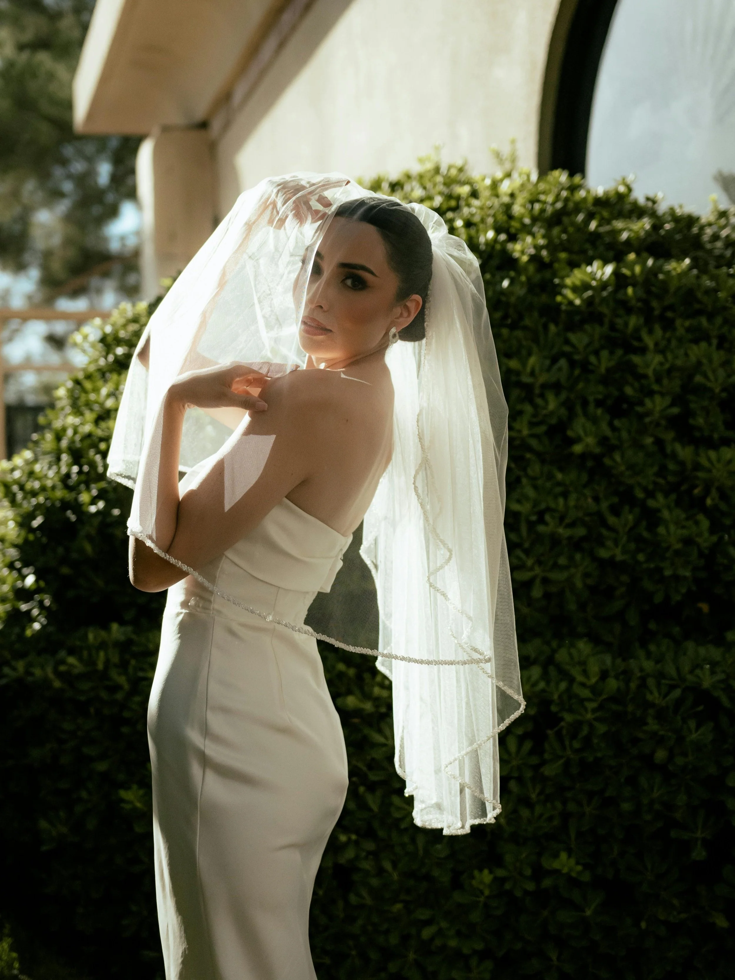 A woman in a white wedding gown and veil standing outdoors beside a hedge with sunlight illuminating her face and shoulders.