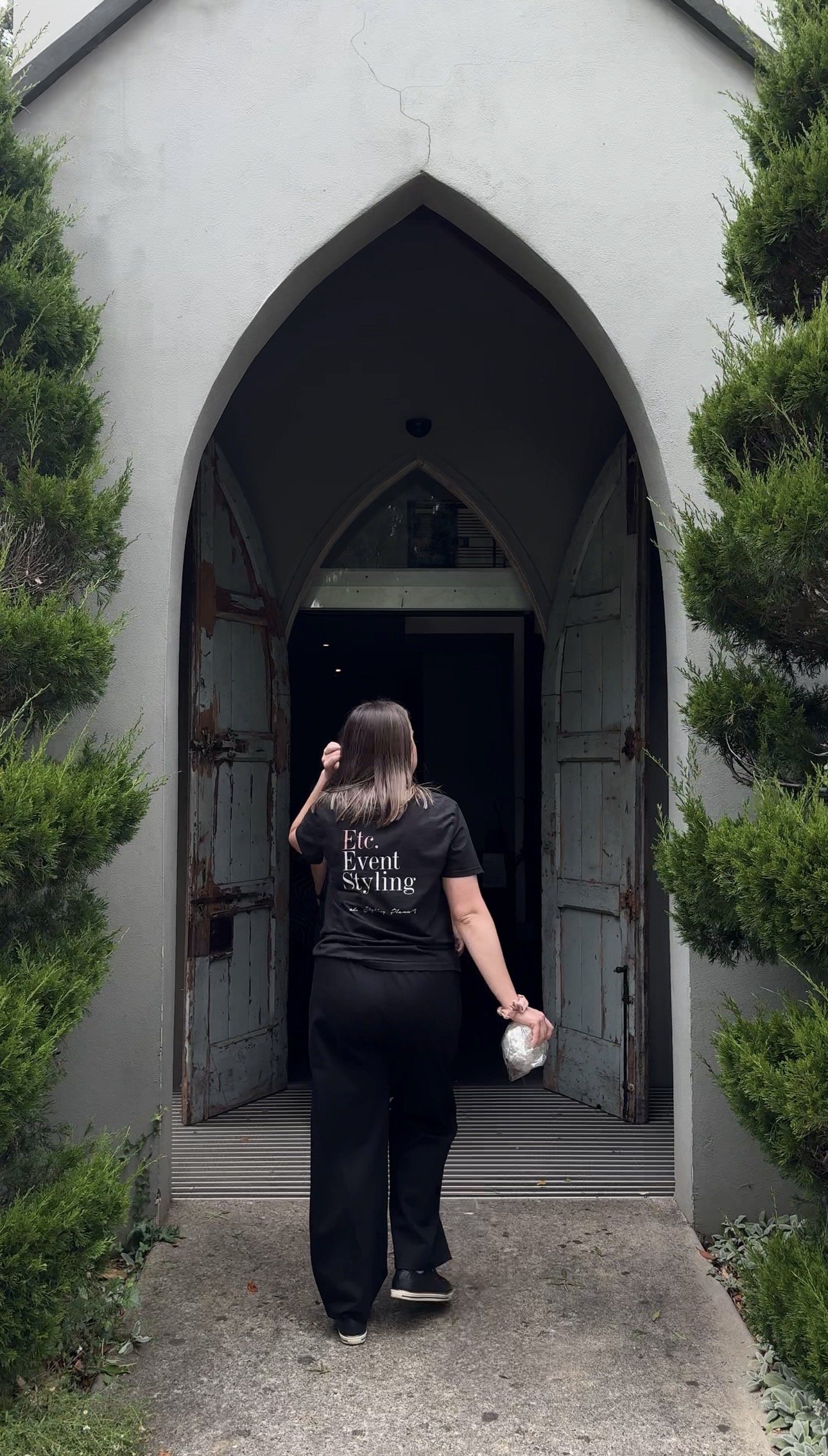 A woman walking toward a historic arched church entrance with rusted double doors, surrounded by greenery.
