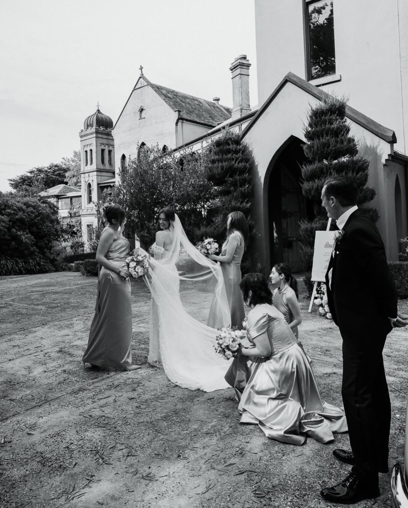 A black and white photo of a wedding ceremony outside a church. The bride and her bridesmaids are standing, with one bridesmaid holding the bride's veil. The groom is standing to the right, watching. An officiant kneels in front, holding a bouquet, as the ceremony takes place on a gravel pathway surrounded by trees and shrubs.