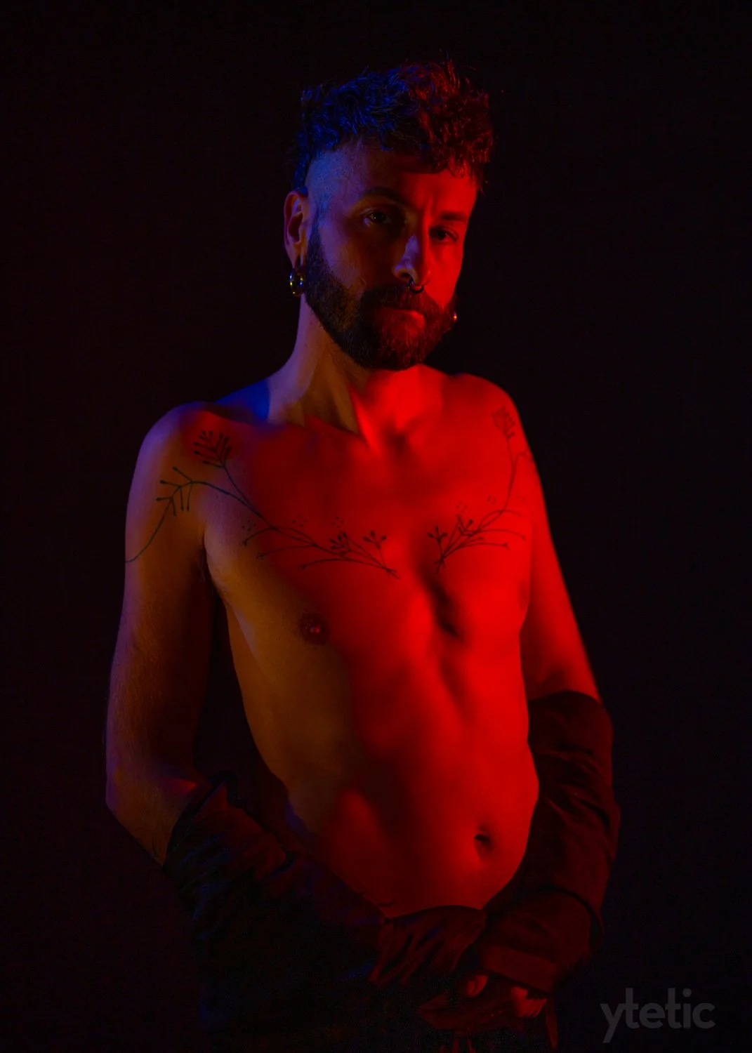 Against a black backdrop, a young man in the process of removing his clothing. He's lit from the right by a red spot and backlit from the left with a blue.