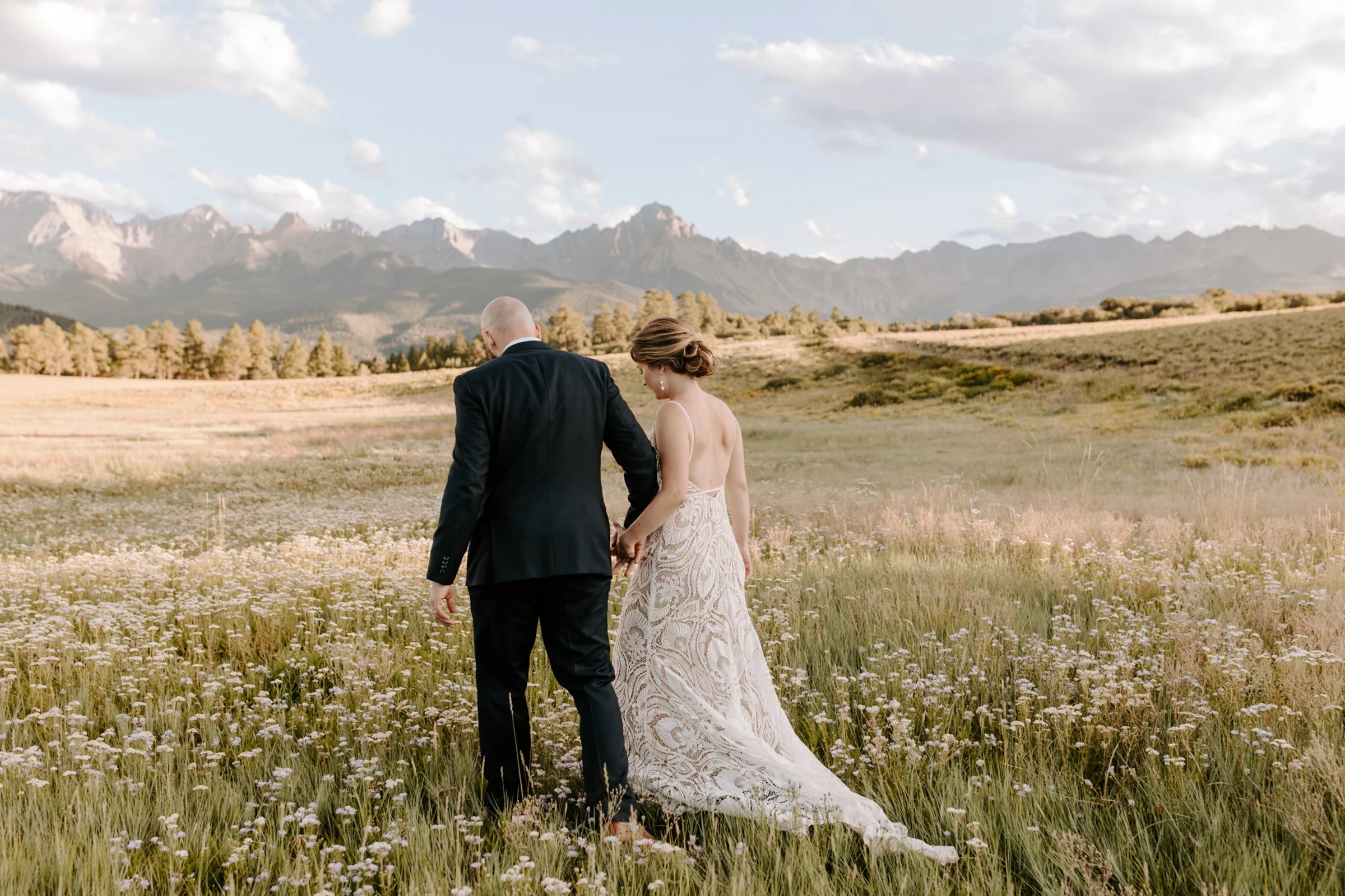 A bride and groom walking hand in hand through a field of wildflowers with mountains in the background.