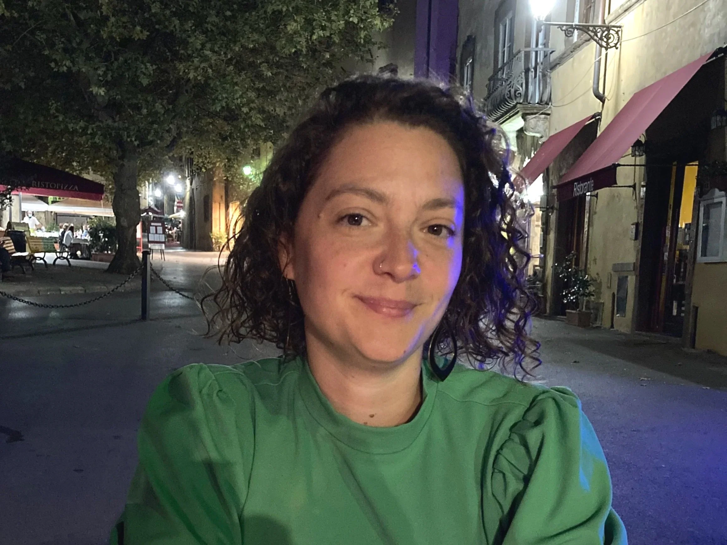 Nighttime street scene with a woman taking a selfie, with lit shops, trees, and empty benches in the background.