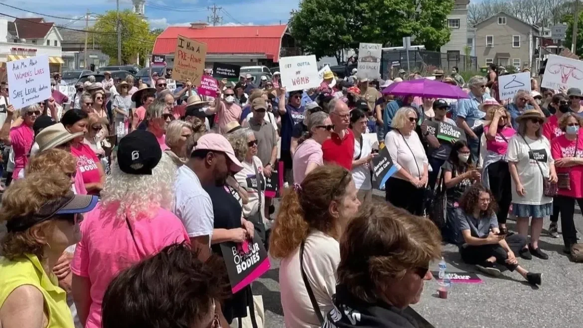 A large group of advocates gathered for a protest, holding signs on a street with trees and buildings in the background. Some signs read 'OUTTA MY WOMB' and 'WE WON'T GO BACK'. Many participants are wearing pink, some with hats.